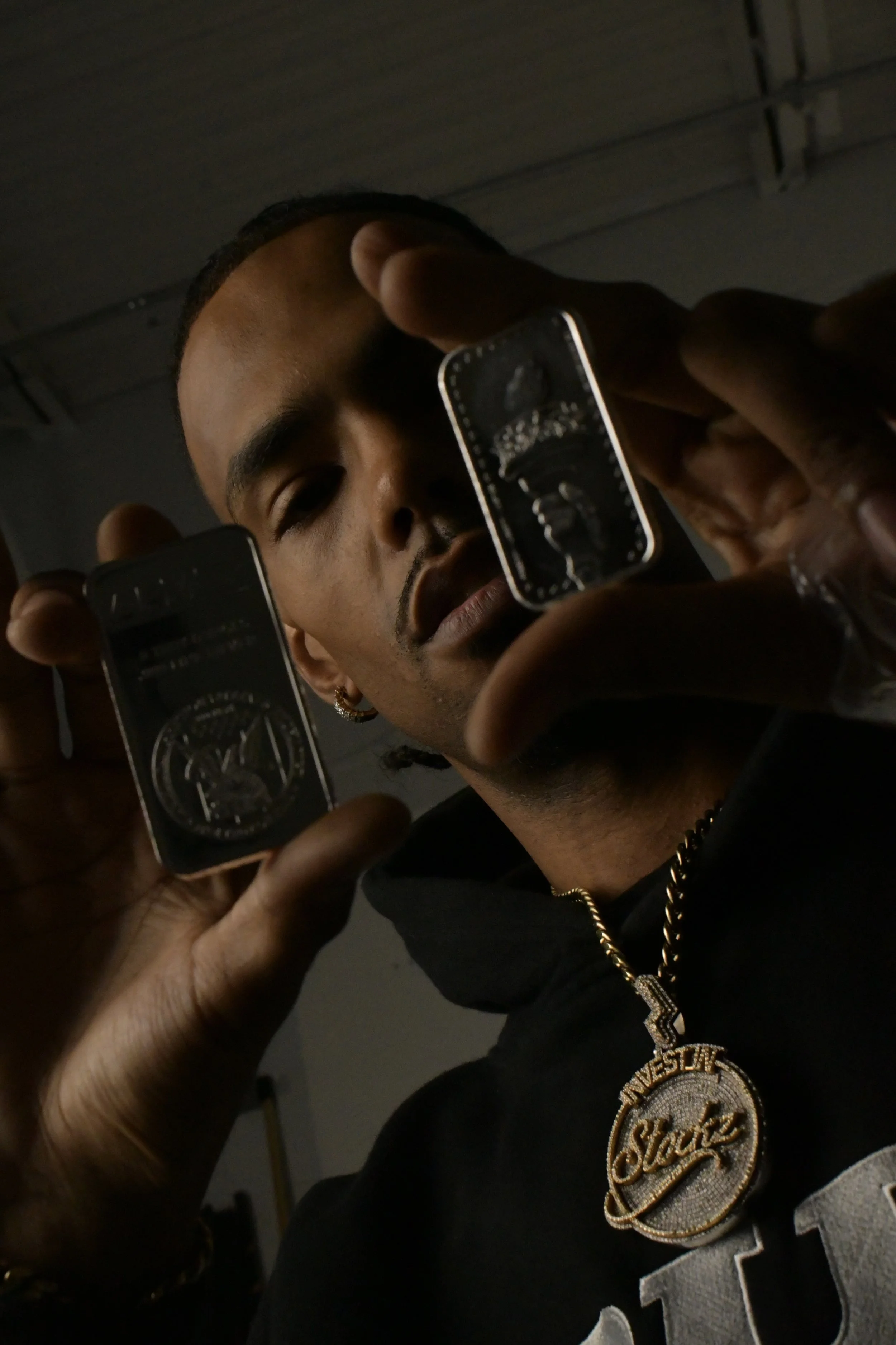 A man with a gold chain and earrings holding two smartphones with black screens, taking a selfie indoors.