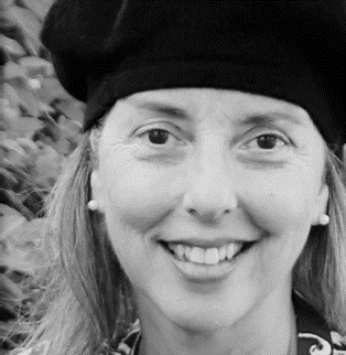 A woman smiling outdoors wearing a black beanie and earrings, with trees in the background.