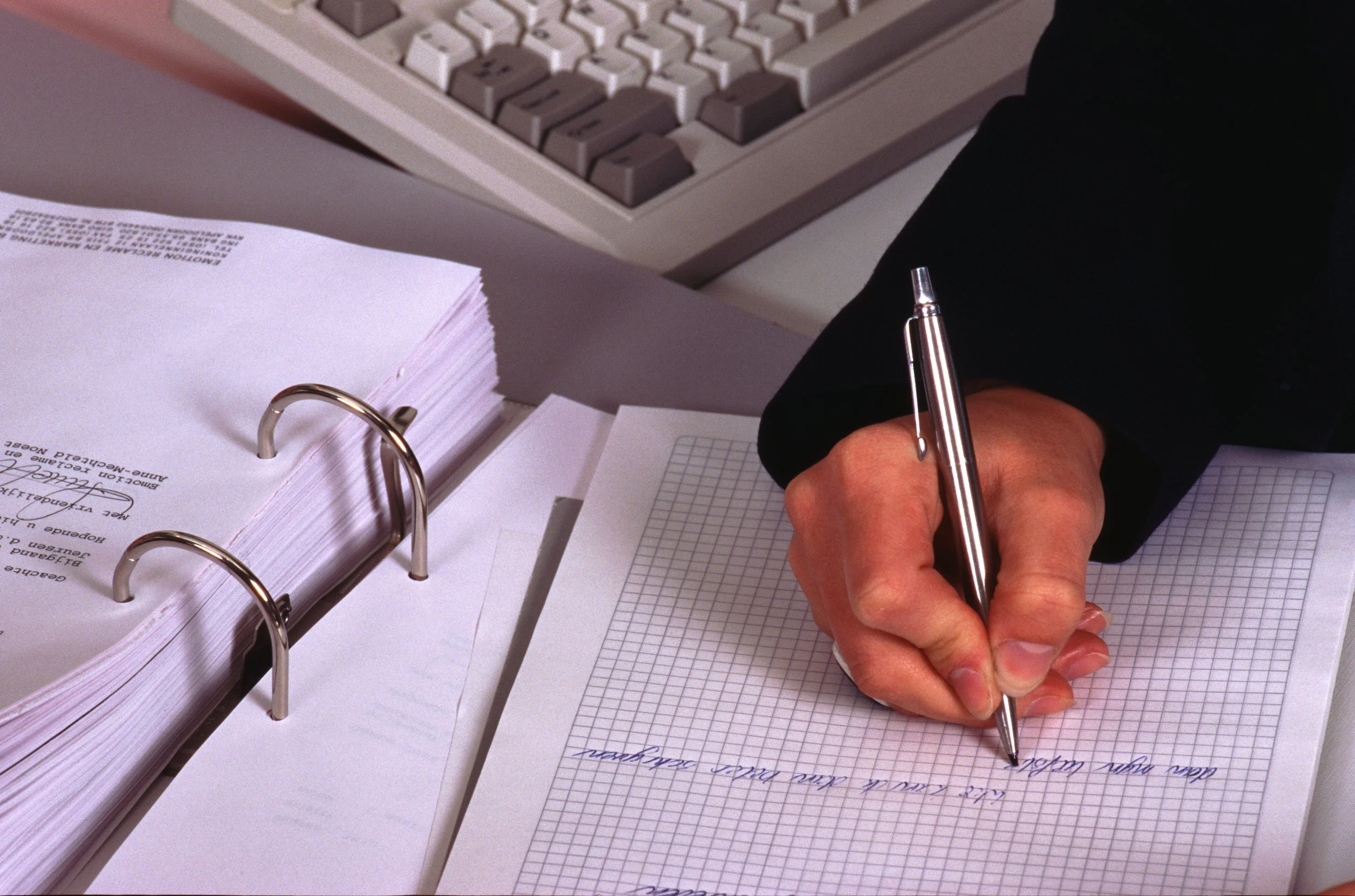 Close-up of a hand writing on graph paper with a silver pen at a desk with a computer keyboard and a binder filled with printed documents.