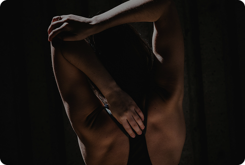 A person with dark hair poses with arms above head, partially covering their chest, in a dark setting with dramatic lighting.