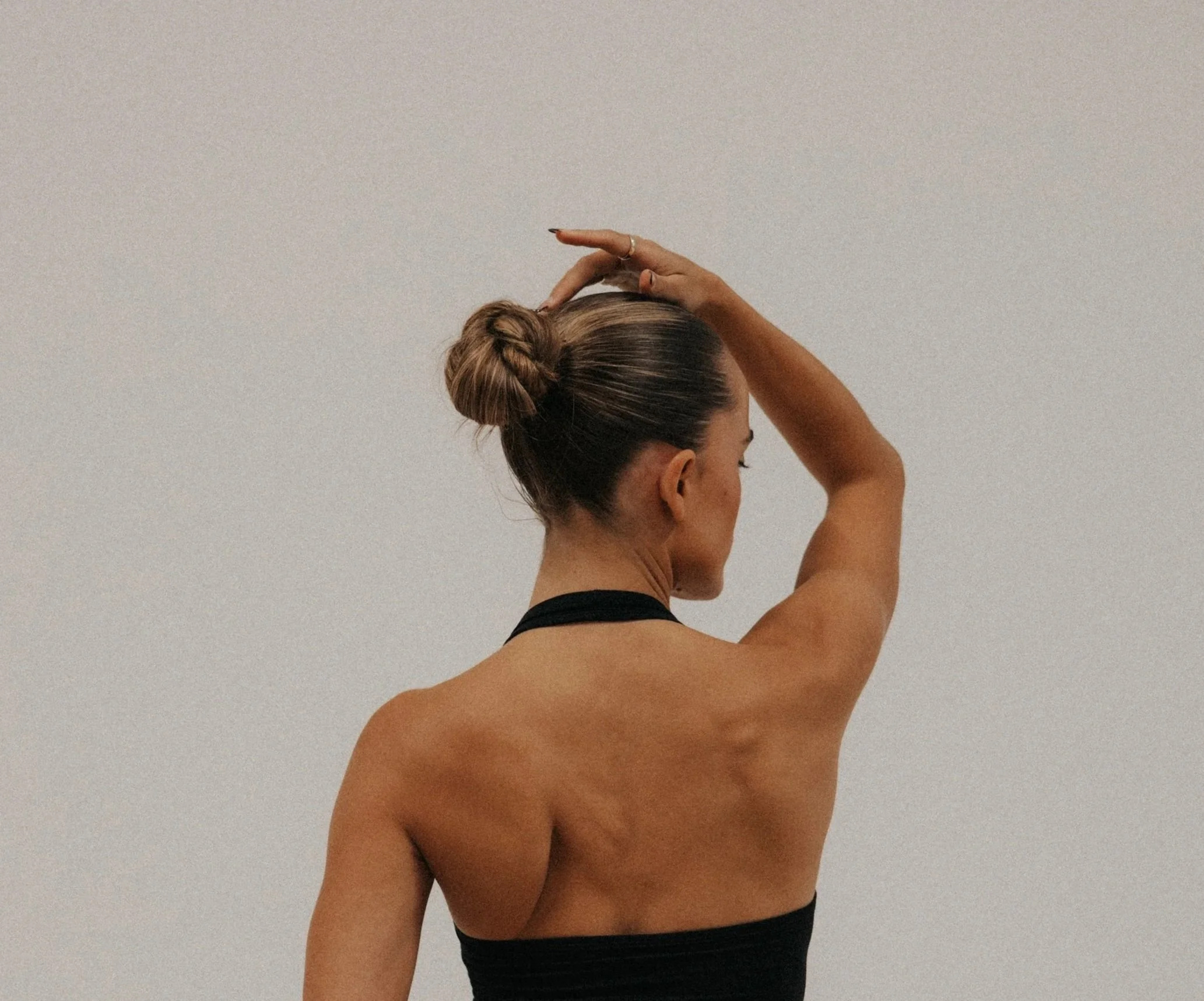 Back view of a woman with a bun hairstyle, wearing a black strap top, standing against a plain background, with one arm raised and hand resting on her head.