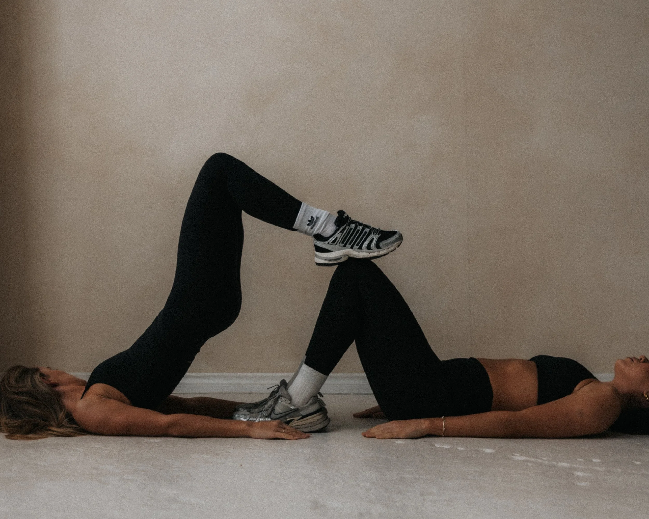 Two women performing a glute bridge exercise together in a room with a beige wall and carpeted floor.