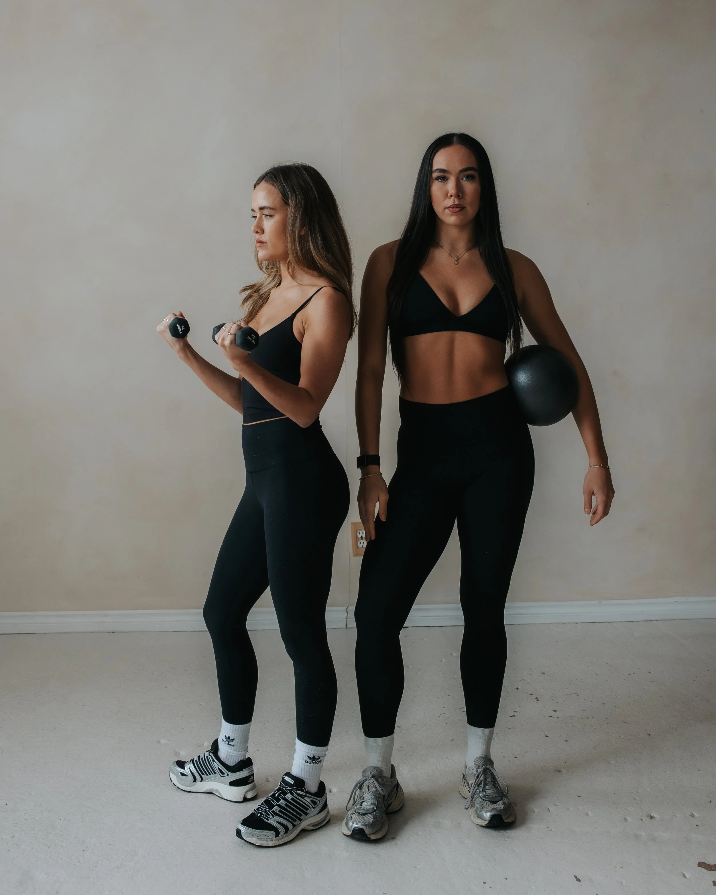 Two women in black workout clothes standing against a plain wall, one holding a dumbbell and the other holding a black medicine ball, both showing a fitness pose.