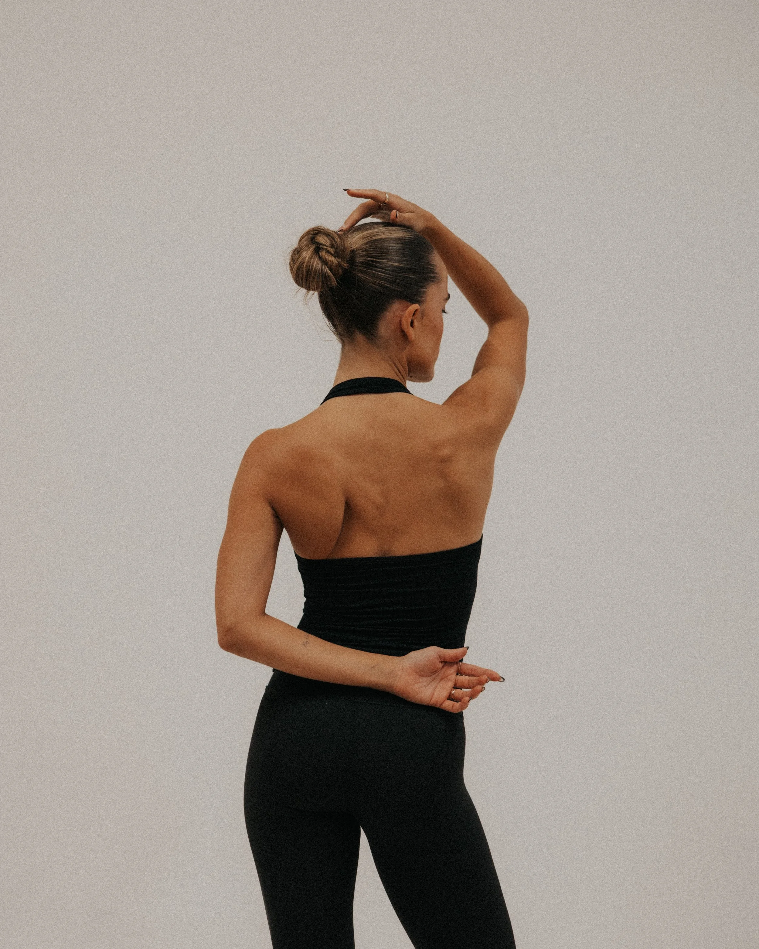A woman with a bun hairstyle practicing yoga or stretching against a plain white wall, wearing a black sleeveless top and black leggings.