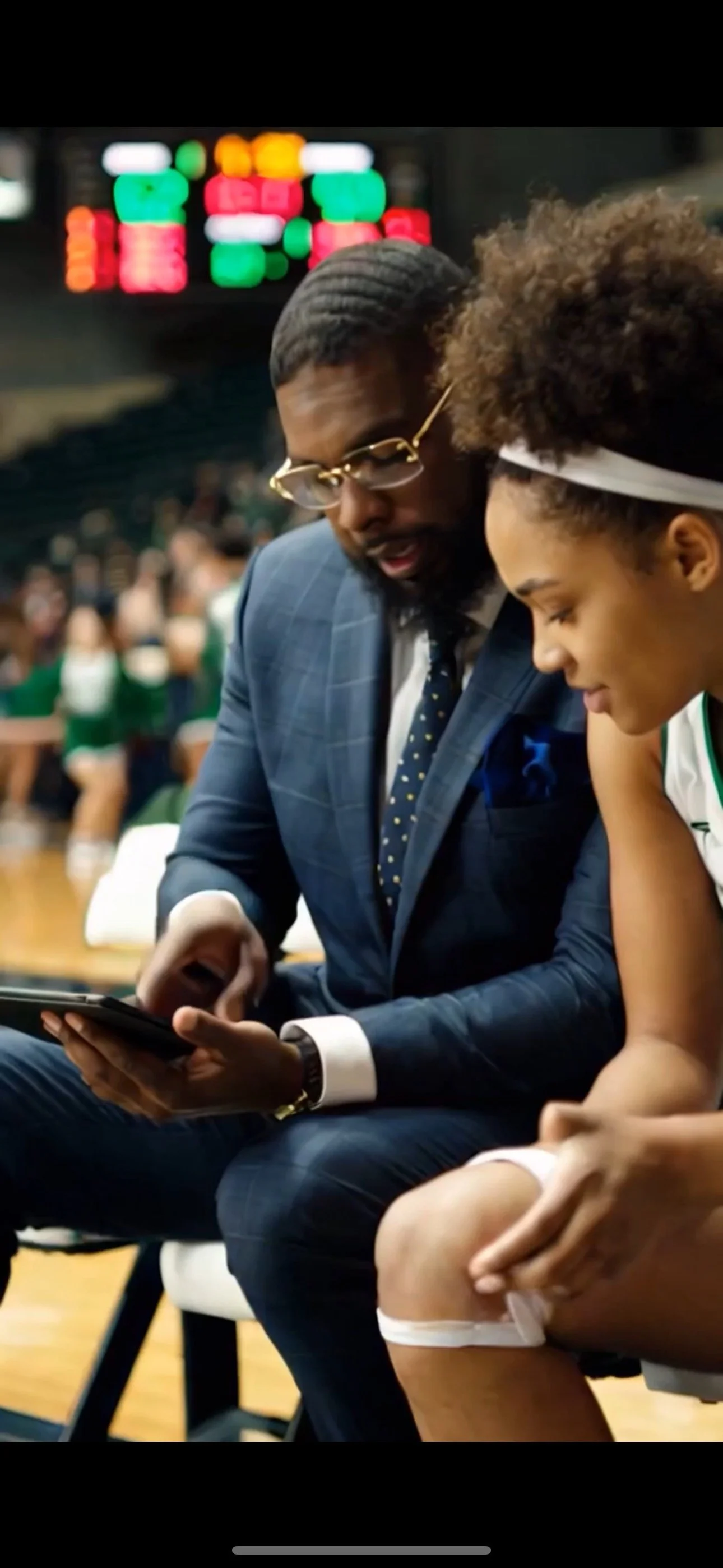 A coach in a blue plaid suit and glasses sitting next to a female basketball player in a white jersey with green accents, both looking at a smartphone during a game, with a scoreboard and spectators in the background.