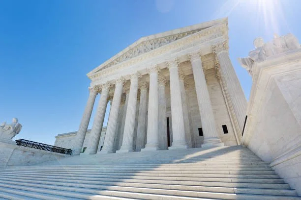 The U.S. Supreme Court building with tall white columns, stairs leading up to the entrance, and a clear blue sky with sunlight.