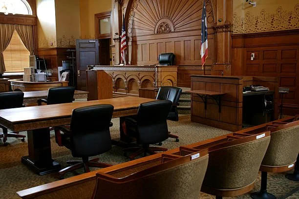 Interior of a courtroom with wooden paneling, a judge's bench, chairs, and American and possibly state flags.