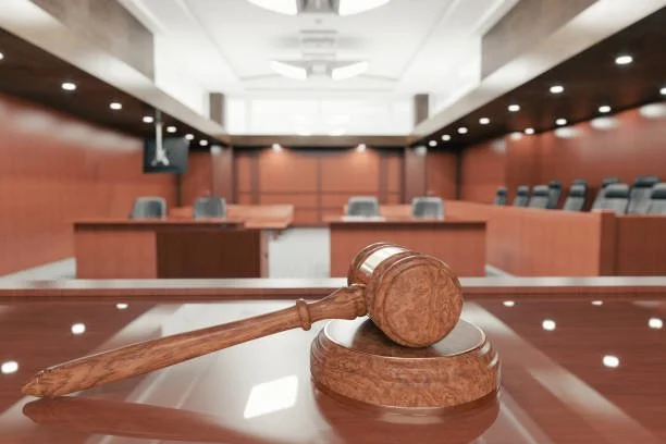 A judge's wooden gavel resting on a sound block in a courtroom with empty chairs and wood-paneled walls.