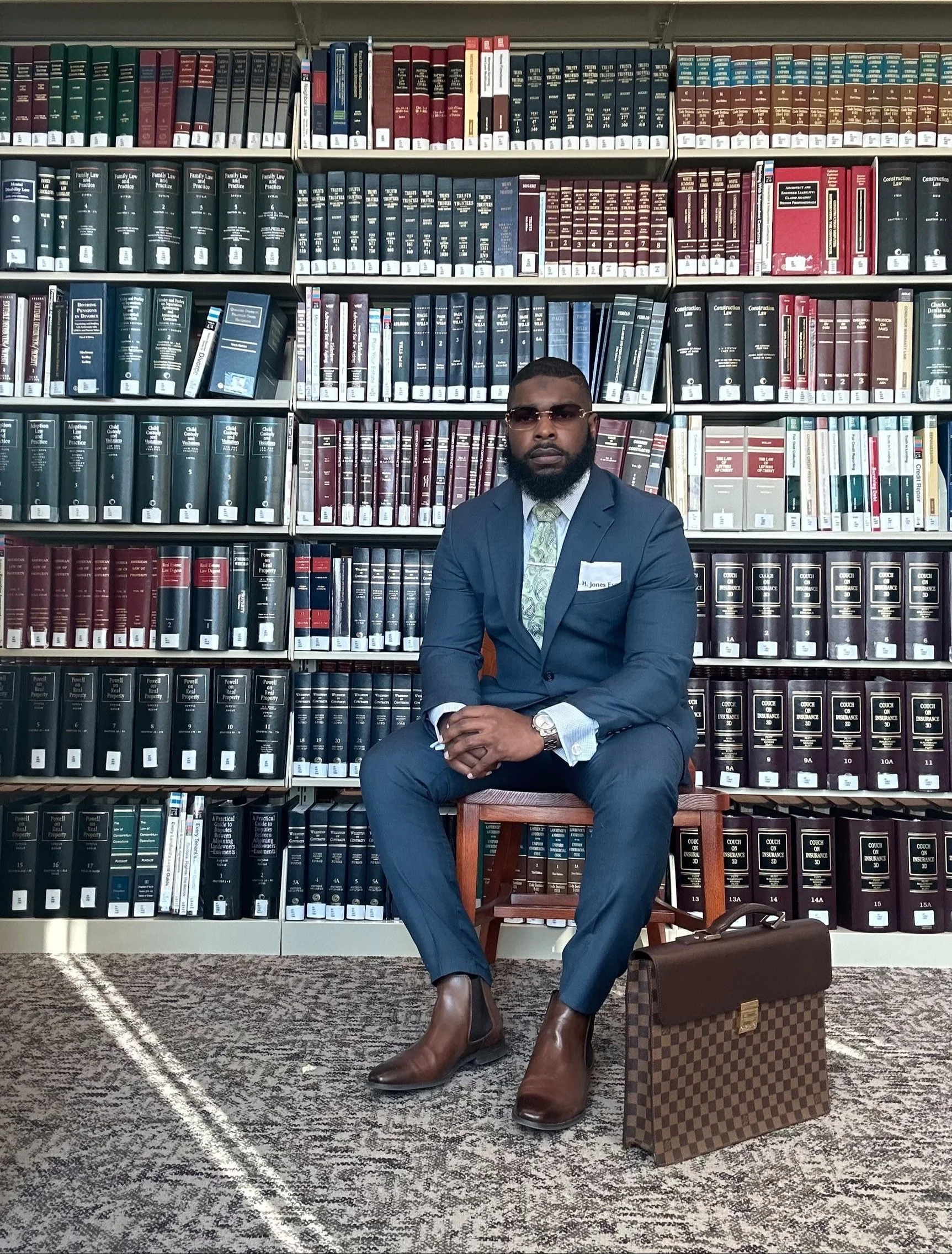 A man dressed in a blue suit, sitting on a wooden chair in front of a bookshelf filled with legal books, holding his hands together. He is wearing sunglasses, a light-colored shirt, a patterned tie, a watch, and has a briefcase placed on the floor beside him.