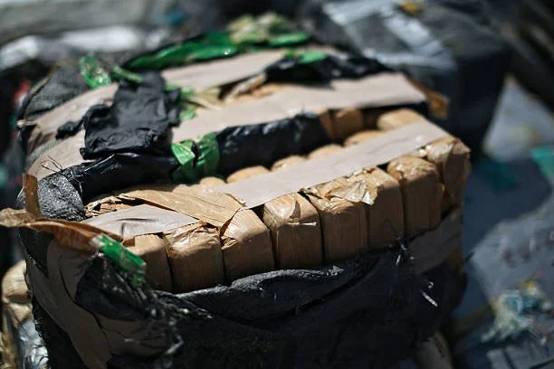A cardboard box wrapped with black and green plastic tape, surrounded by black plastic bags or coverings.