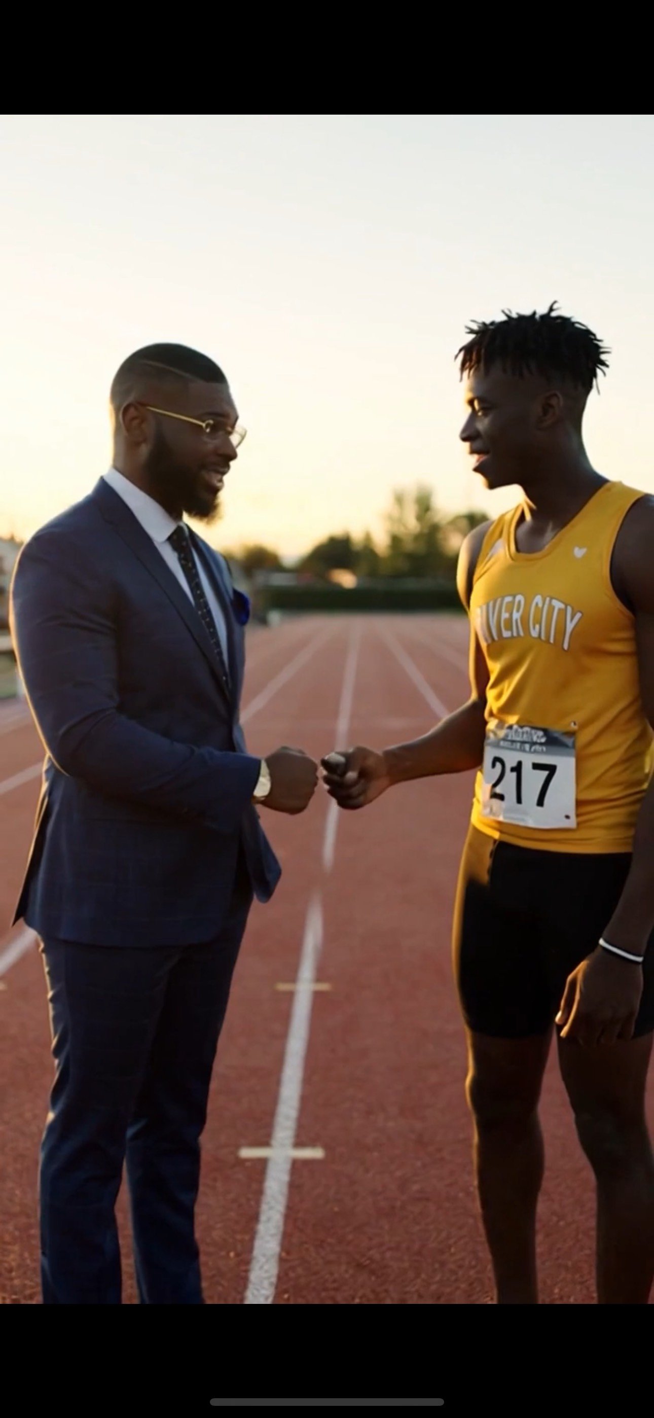 A man in a suit and a runner in a yellow sports uniform shake hands on a track at sunset.