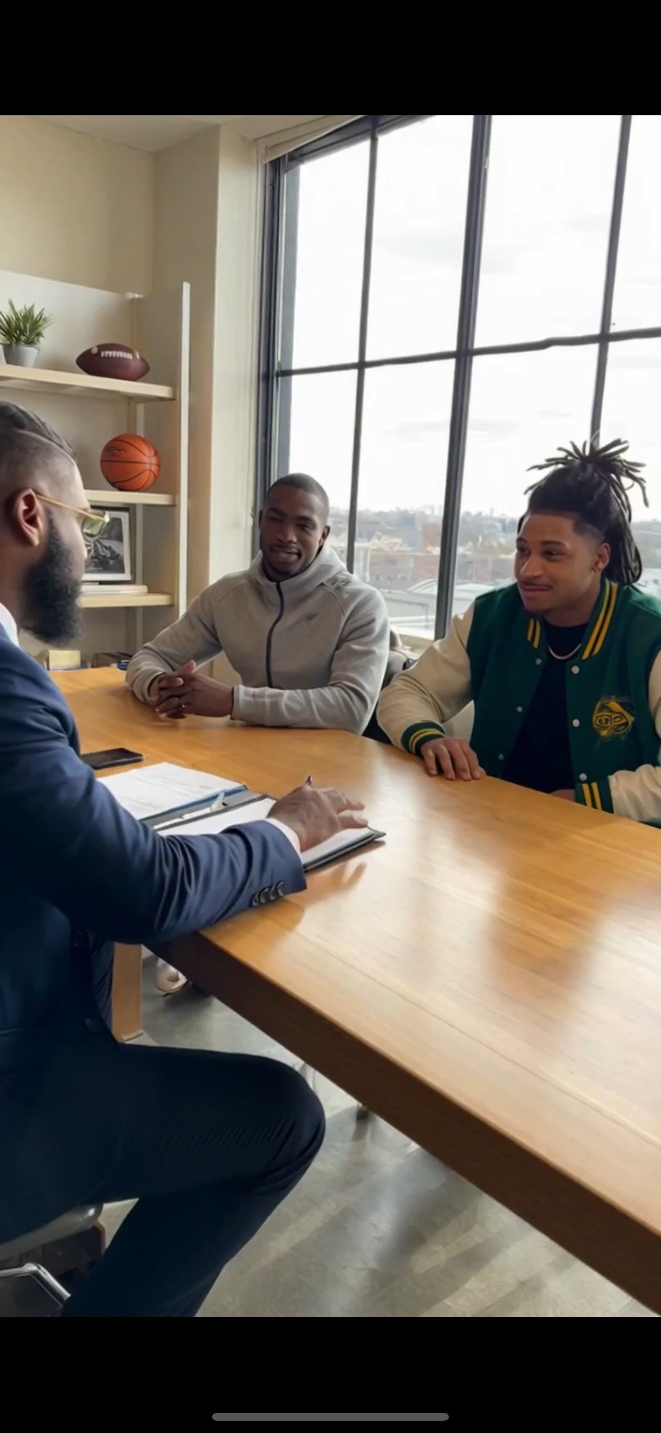 Three men having a discussion at a wooden table in a modern office with large window and sports memorabilia on shelf, including a basketball and a football.