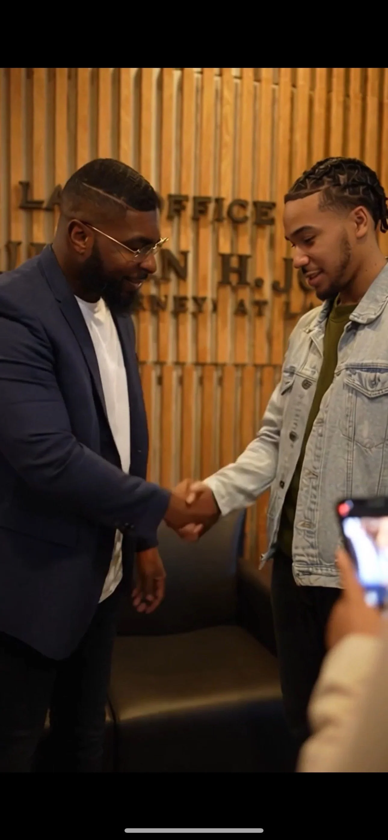 Two men shaking hands in a room with wooden wall decor. One man is wearing a navy blazer and glasses, the other in a light denim jacket, smiling at each other.