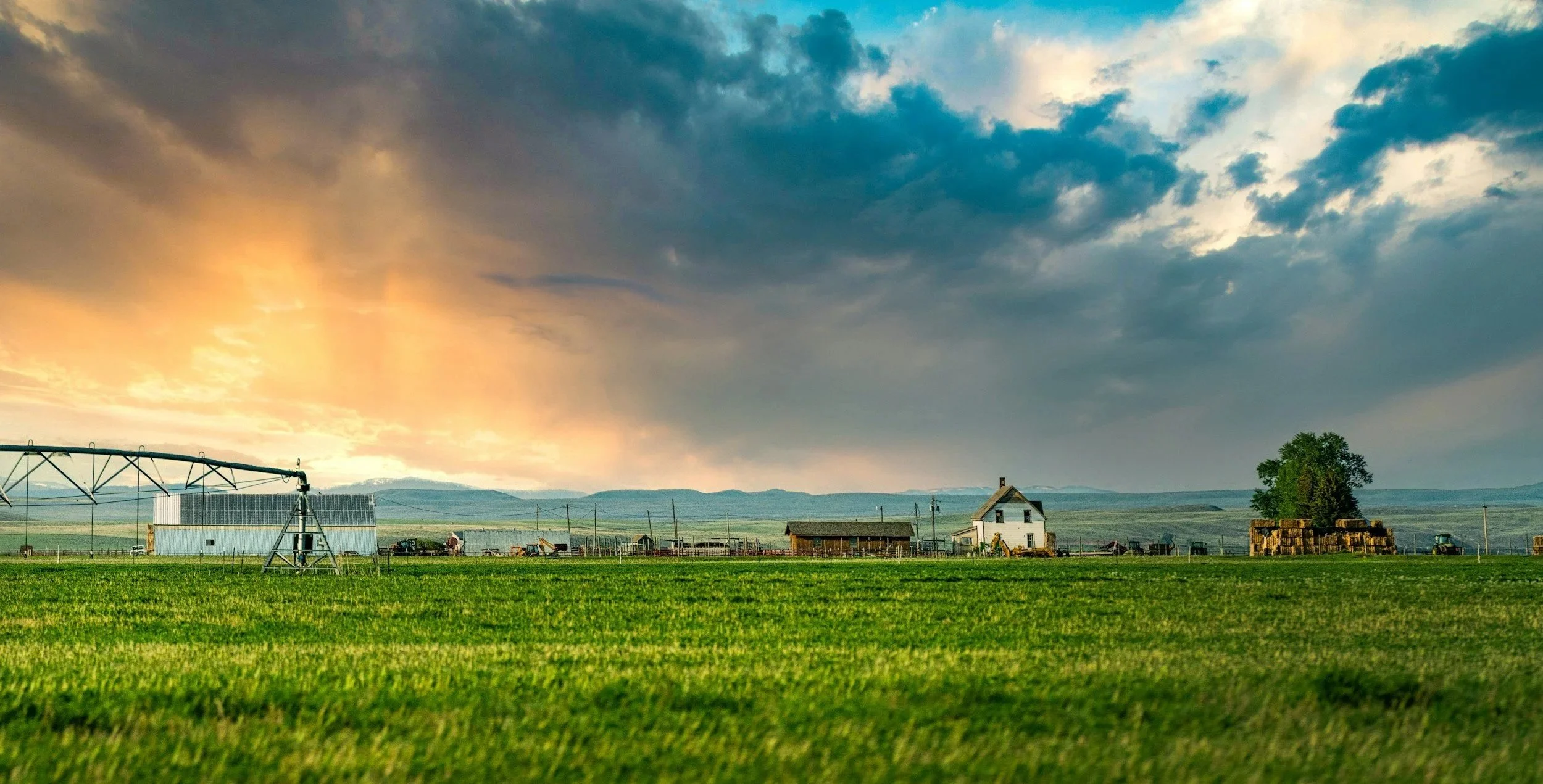 A rural farm landscape during sunset with a green field in the foreground, farm buildings, hay bales, and a large cloudy sky with orange and blue hues.