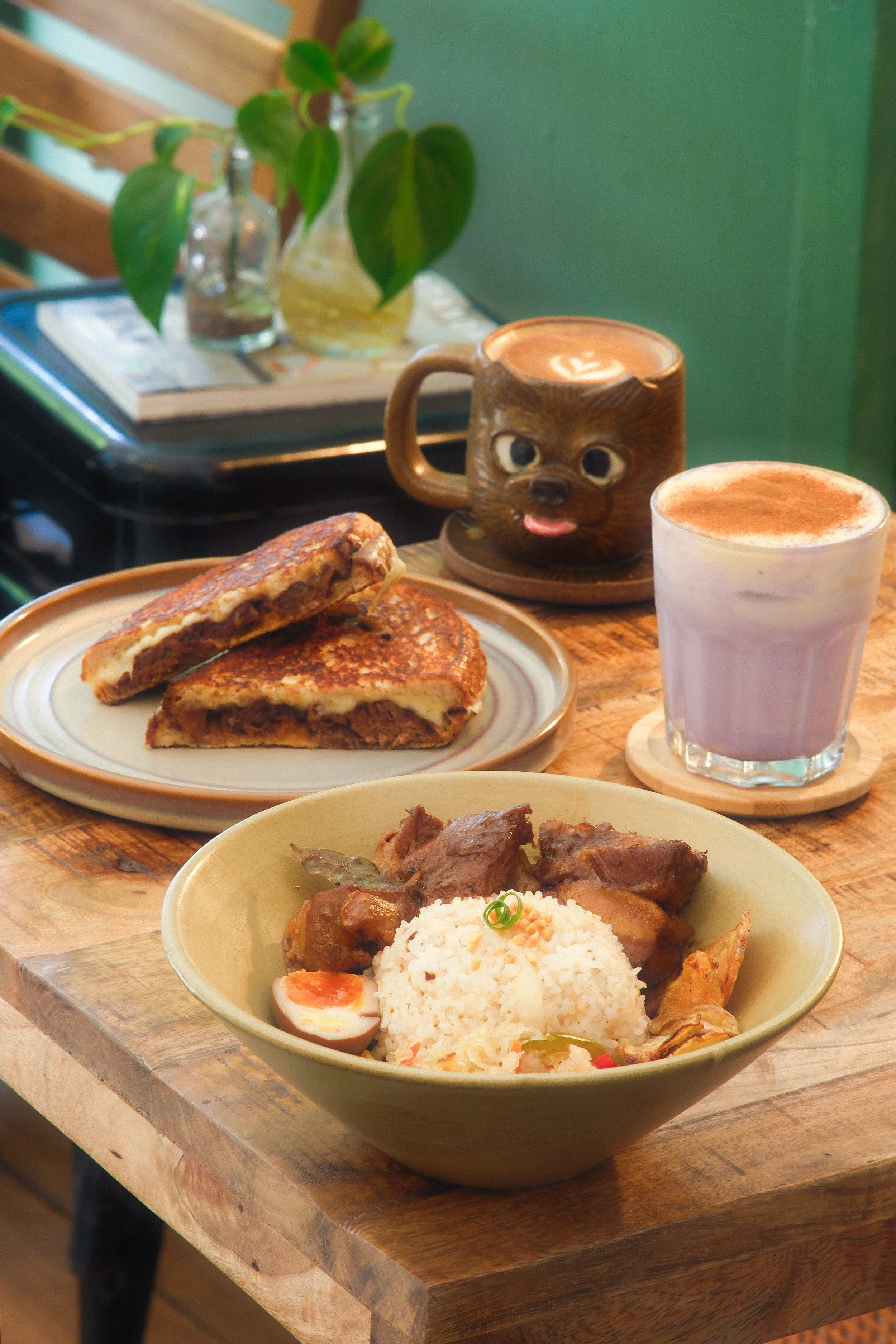 A wooden table with a bowl of rice and meat, a plate with a grilled sandwich, two drinks (one pink latte and one hot chocolate in a bear-shaped mug), and a small plant in the background.