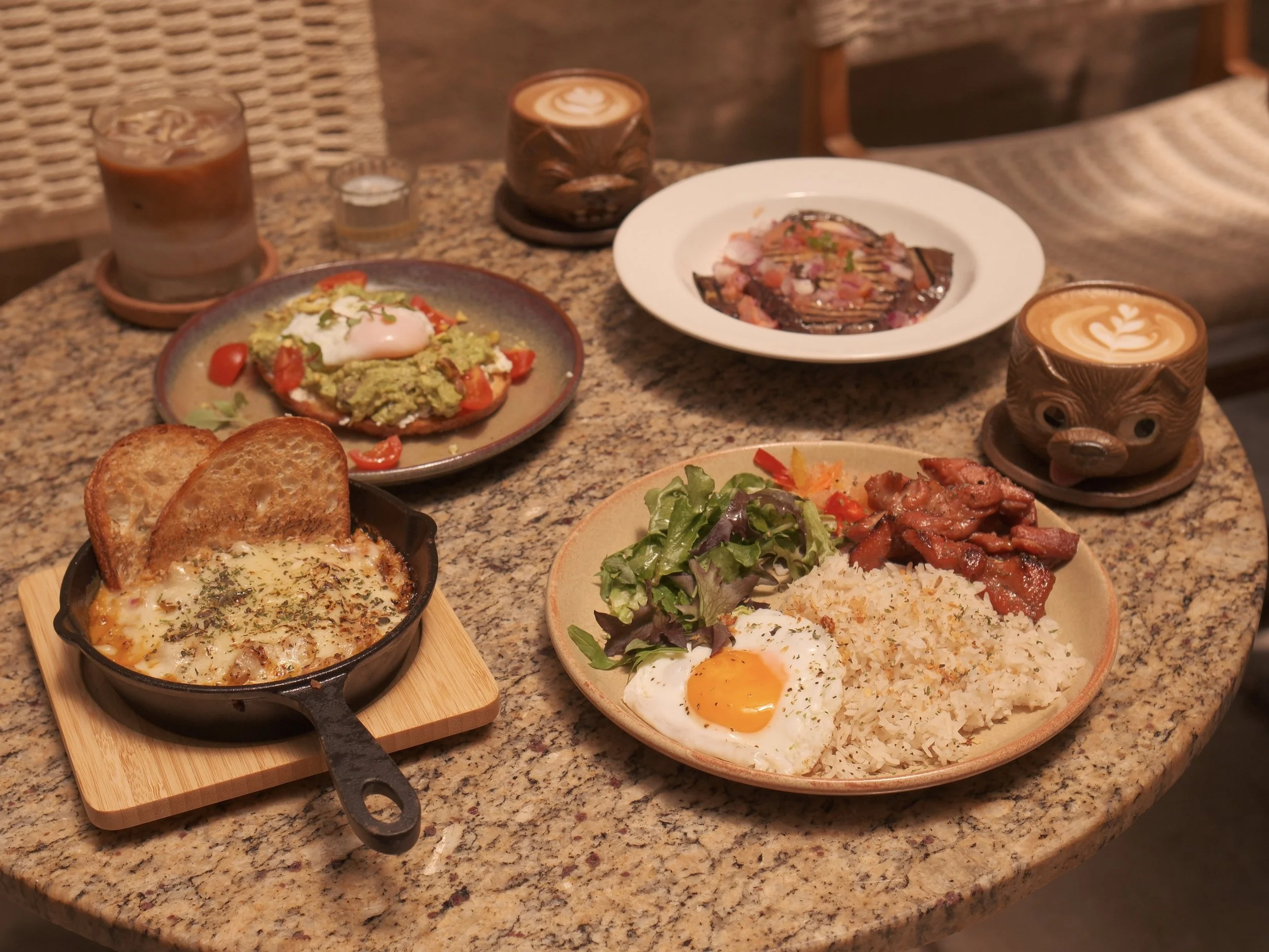 A table with various dishes including a baked dish with toast, a plate of rice with fried egg, bacon, and salad, guacamole with cherry tomatoes and a poached egg, grilled meat with sauce, and two cups of coffee with bear-shaped cups, on a granite tab
