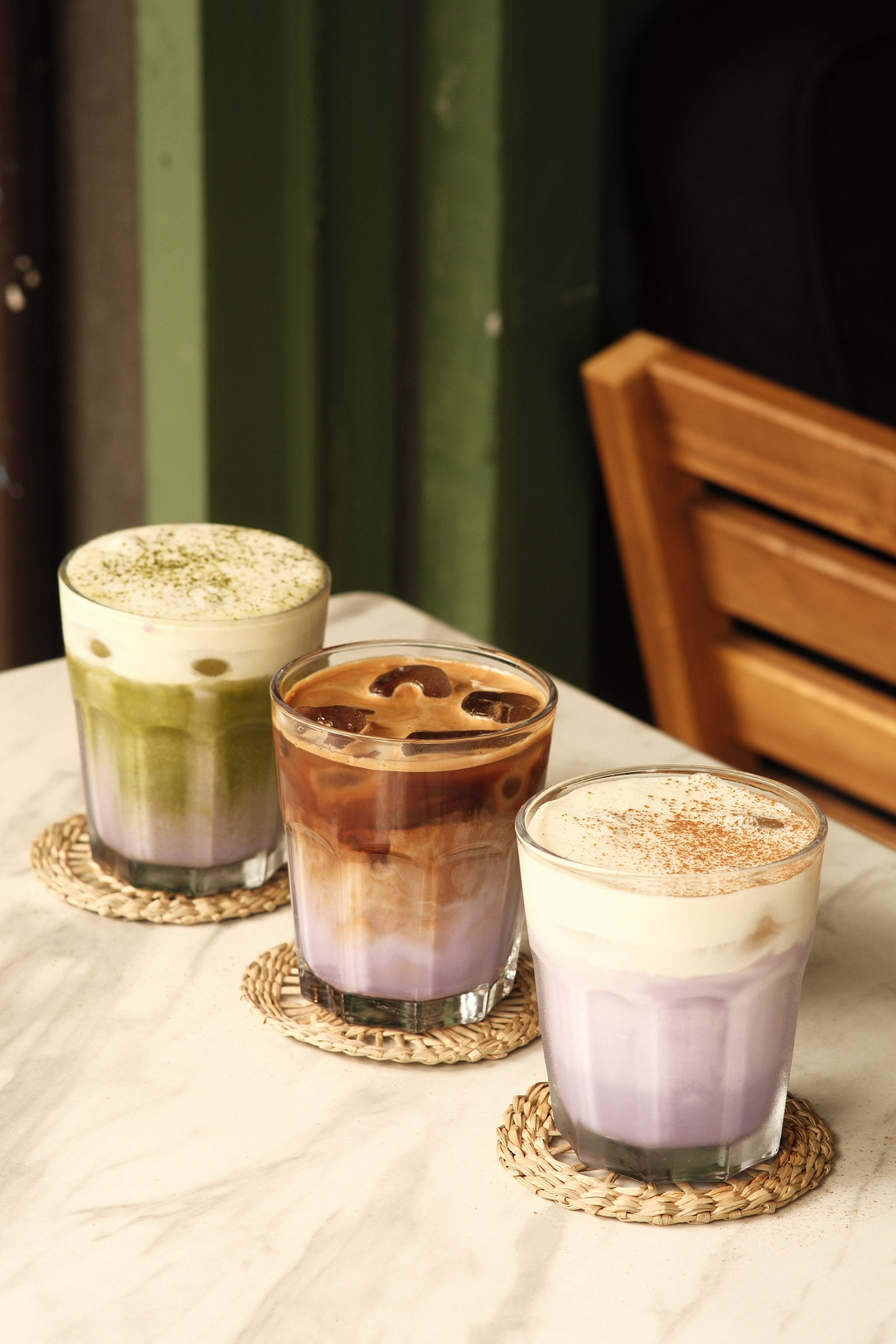 Three colorful layered drinks on a white marble table, each on a woven coaster, with a wooden chair and green wall in the background.
