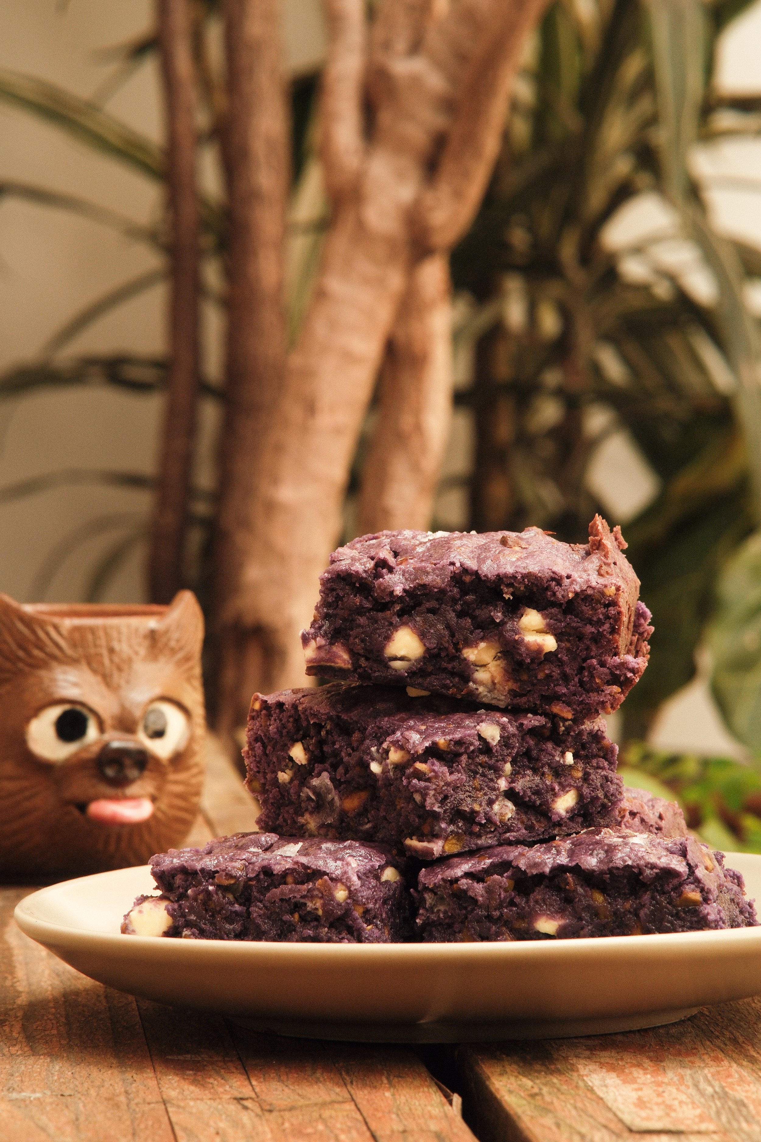 A plate of blueberry dessert squares with nuts, stacked on a wooden surface with plants and a ceramic animal figure in the background.