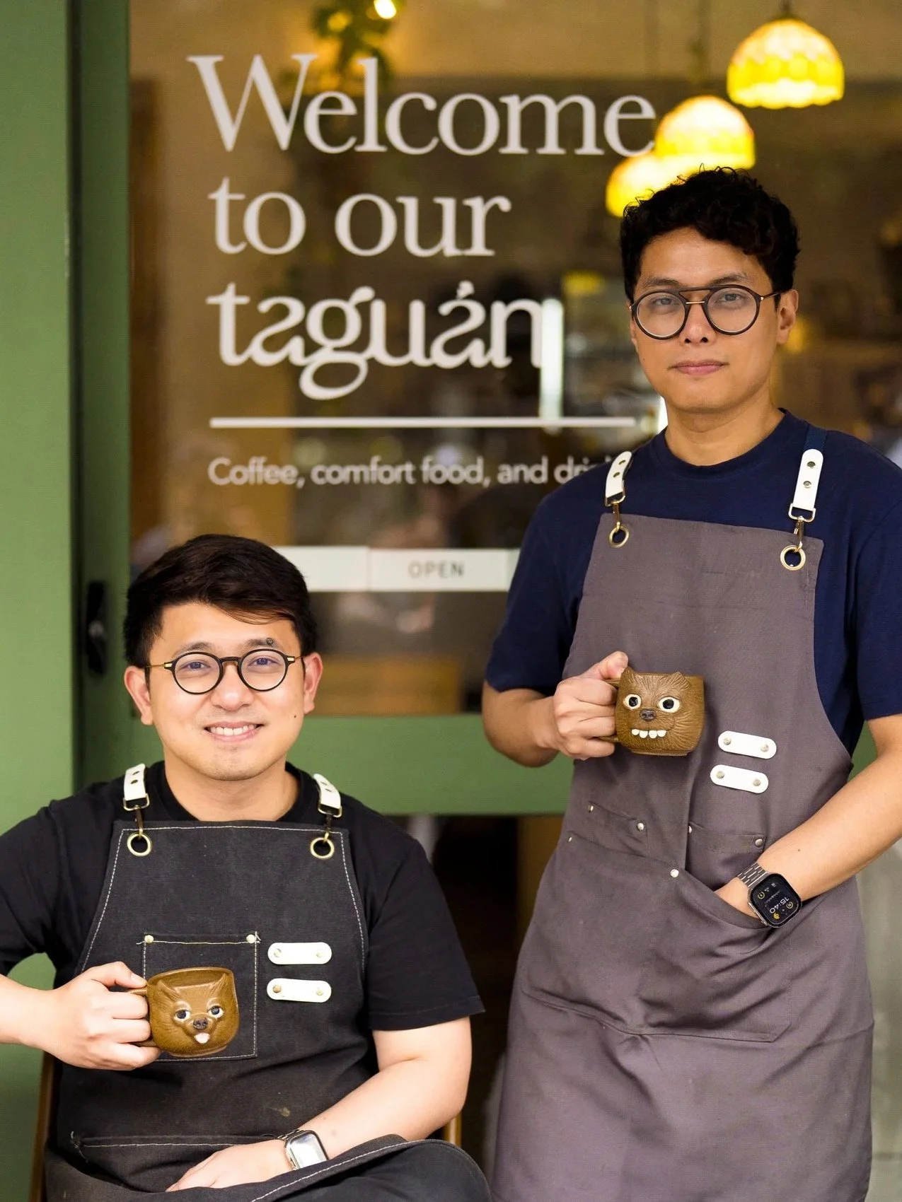 Two men with glasses wearing aprons, holding coffee mugs shaped like a cartoon animal, standing in front of a glass door with a sign that reads, 'Welcome to our taguán,' offering coffee, comfort food, and drinks.