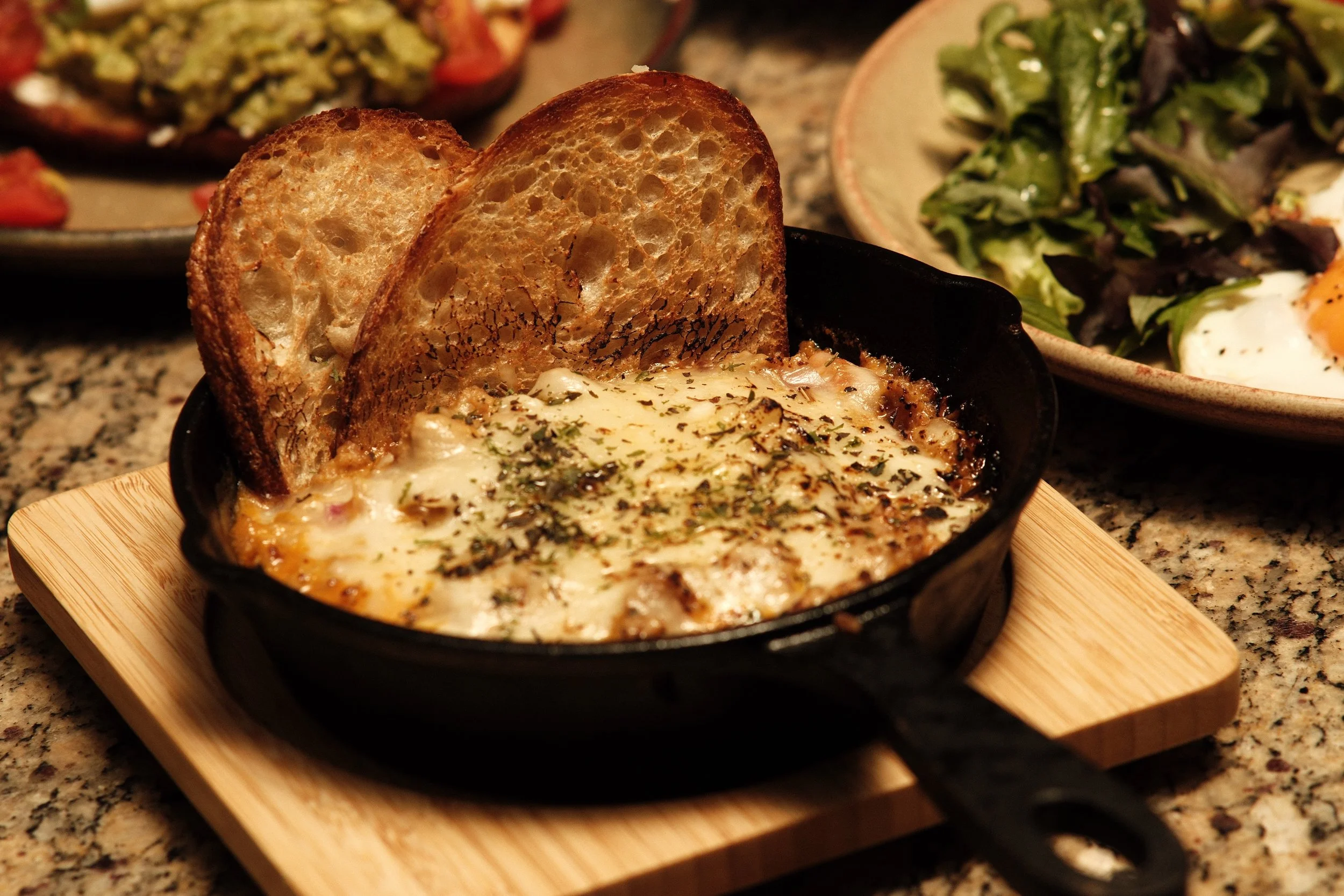A small black skillet of baked cheesy casserole topped with herbs and two toasted bread slices, with salads in the background.