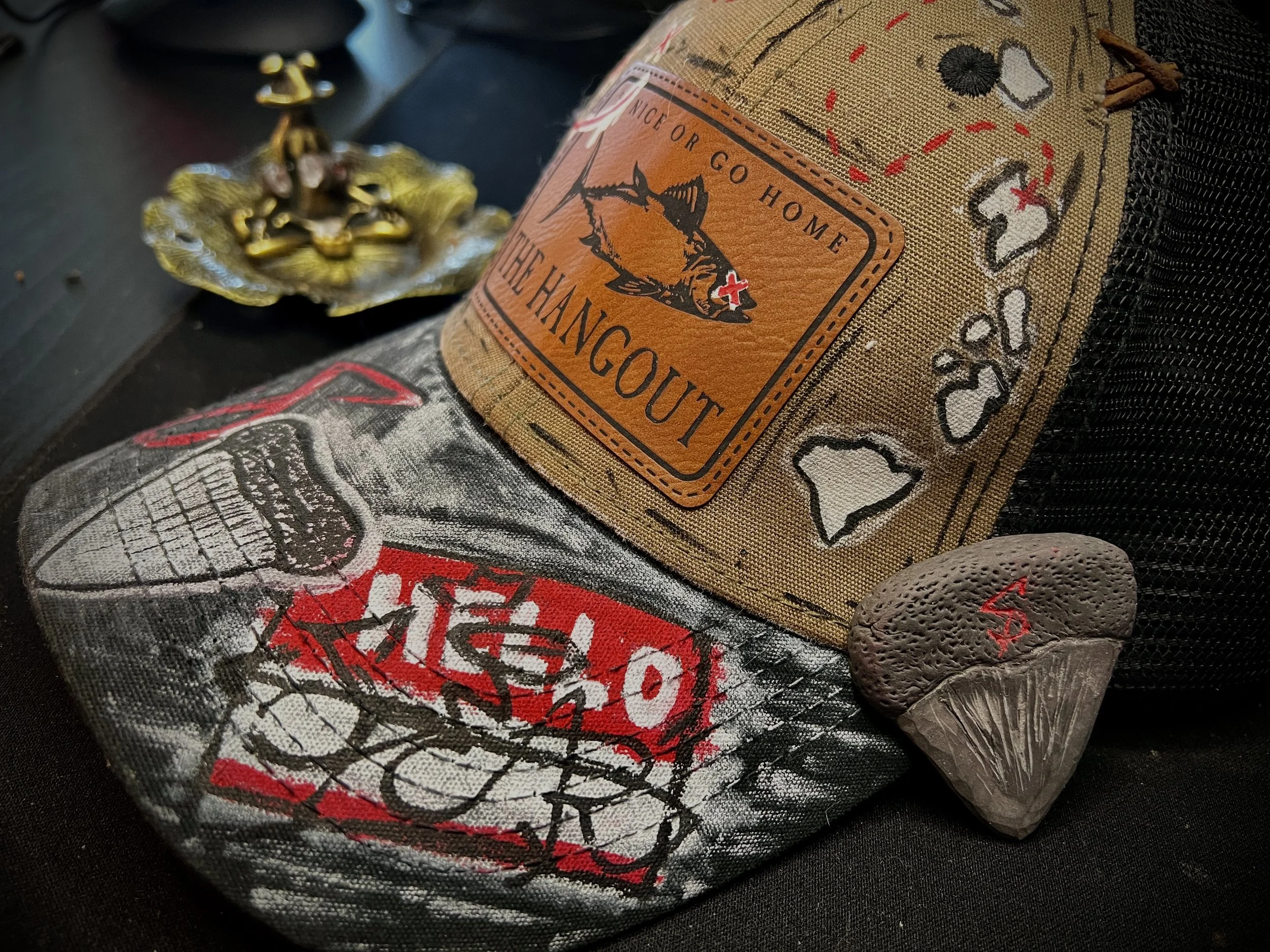 Close-up of a patch with a fish illustration and the words "Go Nice Or Go Home" and "In The Hangout." A fabric with a graphic design, a small gray stone with a red marking, and a brass decorative item are also visible on a dark surface.