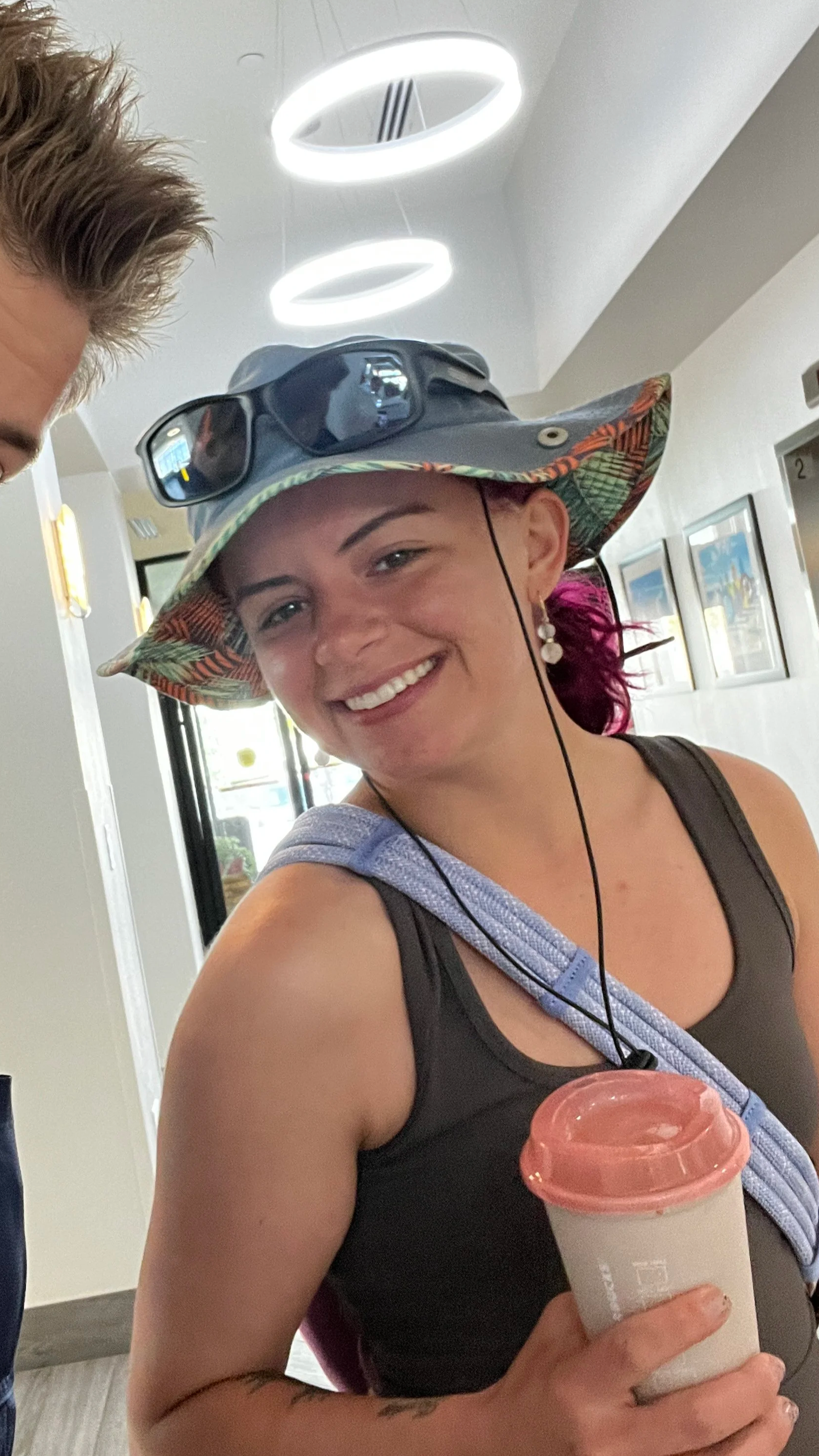 Smiling woman with pink hair wearing a large floral hat, sunglasses pushed up on the hat, and a black tank top, holding a coffee cup, inside a brightly lit indoor space.