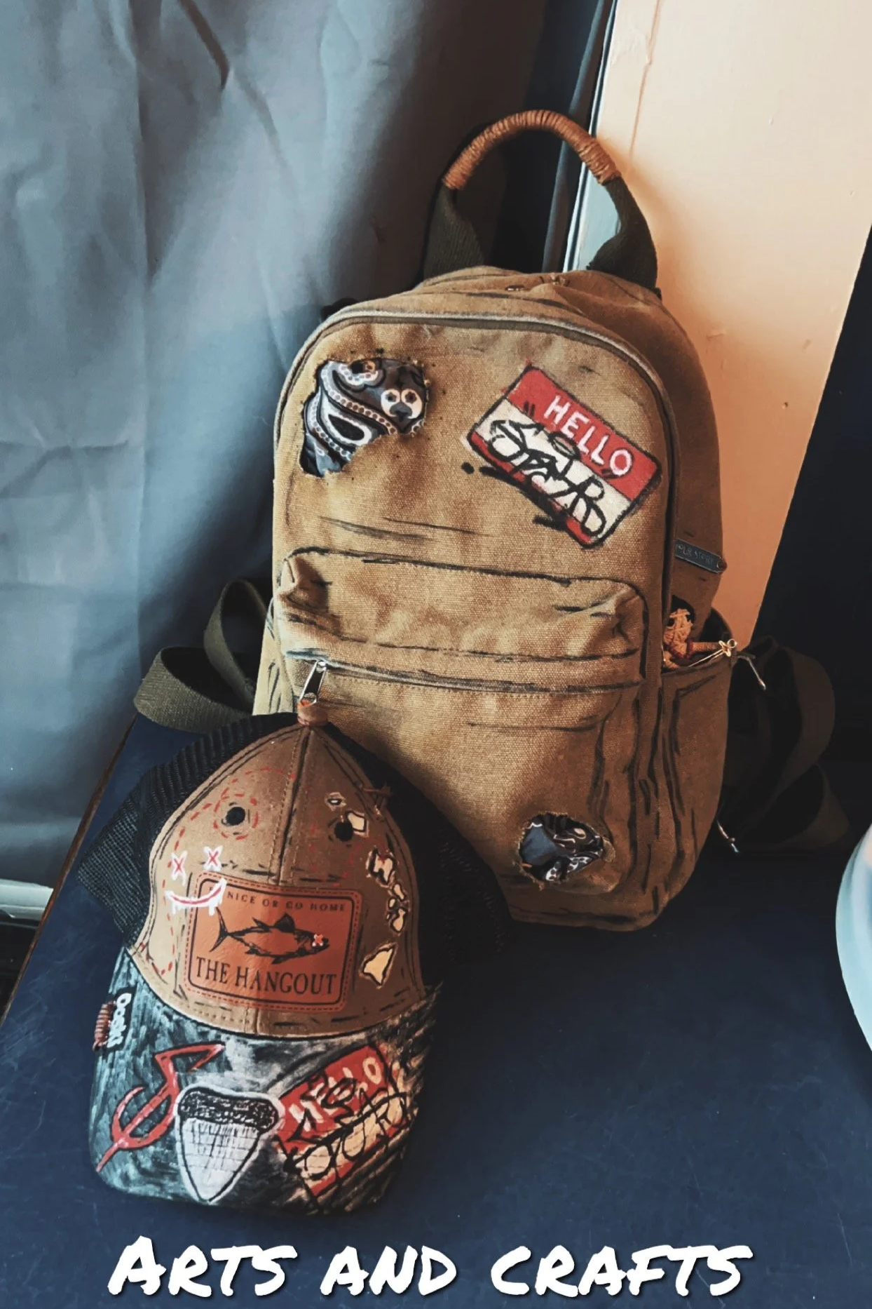 A tan backpack with patches and a cap with patches, displayed on a black surface. The patches on the backpack include one with a snake and another with a Hello Sports logo. The cap has patches with a shark and a speech bubble saying "Nice or Go Home!" and "The Hangout." The text "ARTS AND CRAFTS" is overlaid at the bottom of the image.