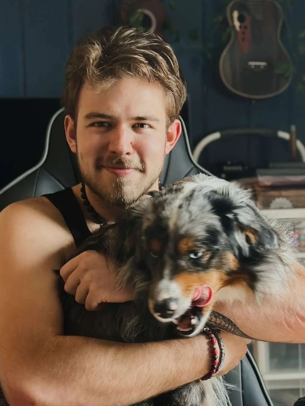 A man with light brown hair and a beard is sitting in a chair, smiling, holding a merle Australian Shepherd dog with blue eyes. The dog is licking its nose, and the man is holding it gently. In the background, there are guitars hanging on a wall.