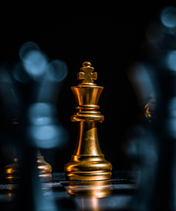 A golden chess queen piece on a reflective surface with blurred blue lights in the background.