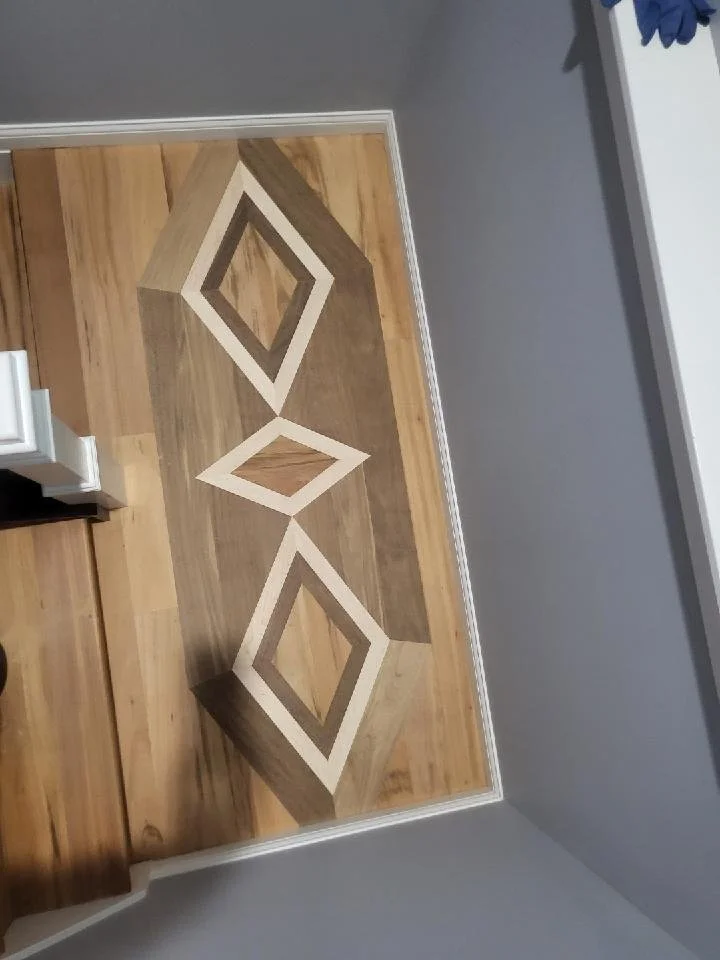 Close-up of a patterned wooden floor with geometric inlays of diamond shapes in front of a gray wall and white trim.