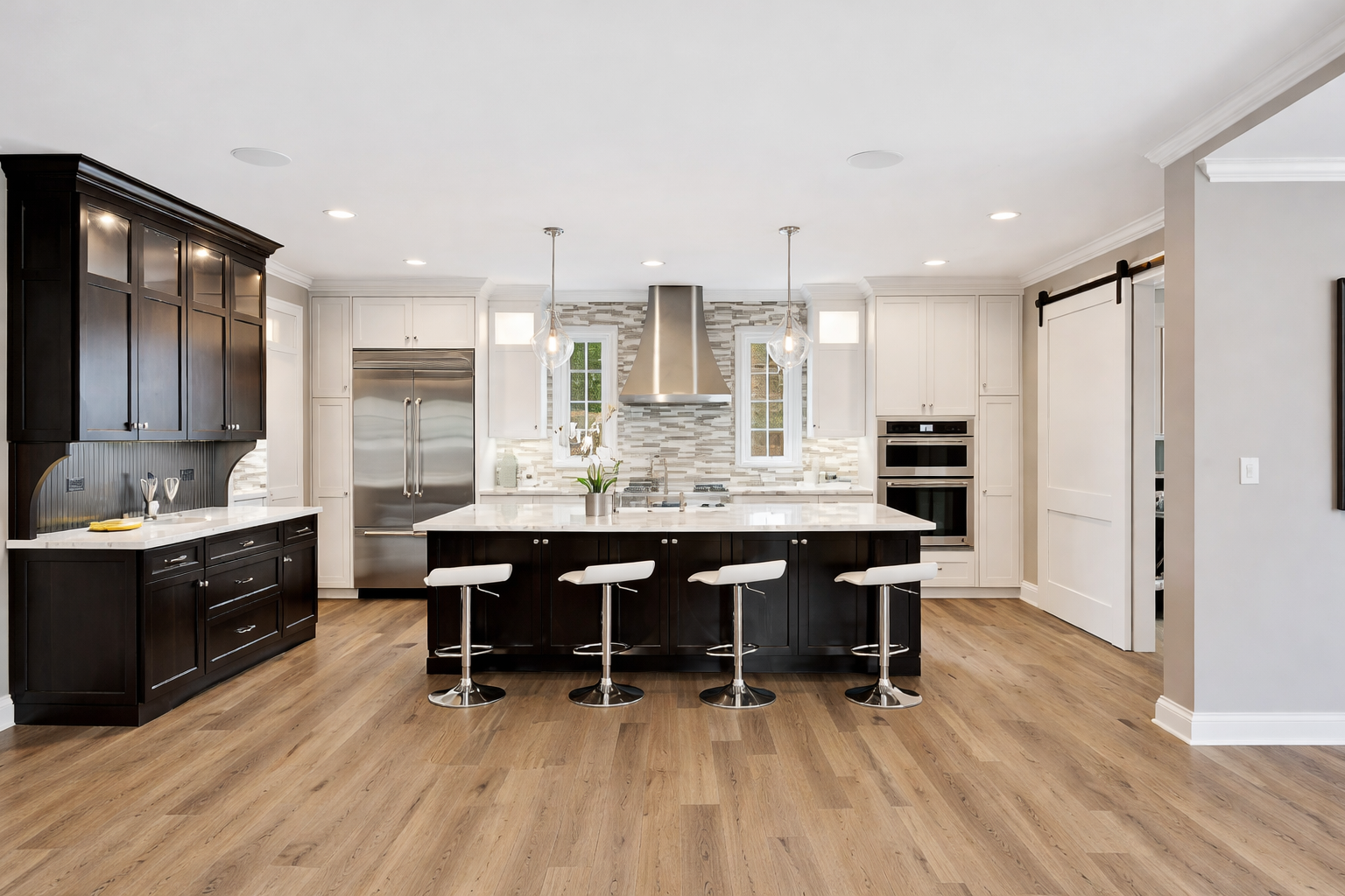 Modern kitchen with black and white cabinetry, marble island with four white barstools, stainless steel appliances, wooden flooring, and a tiled backsplash.