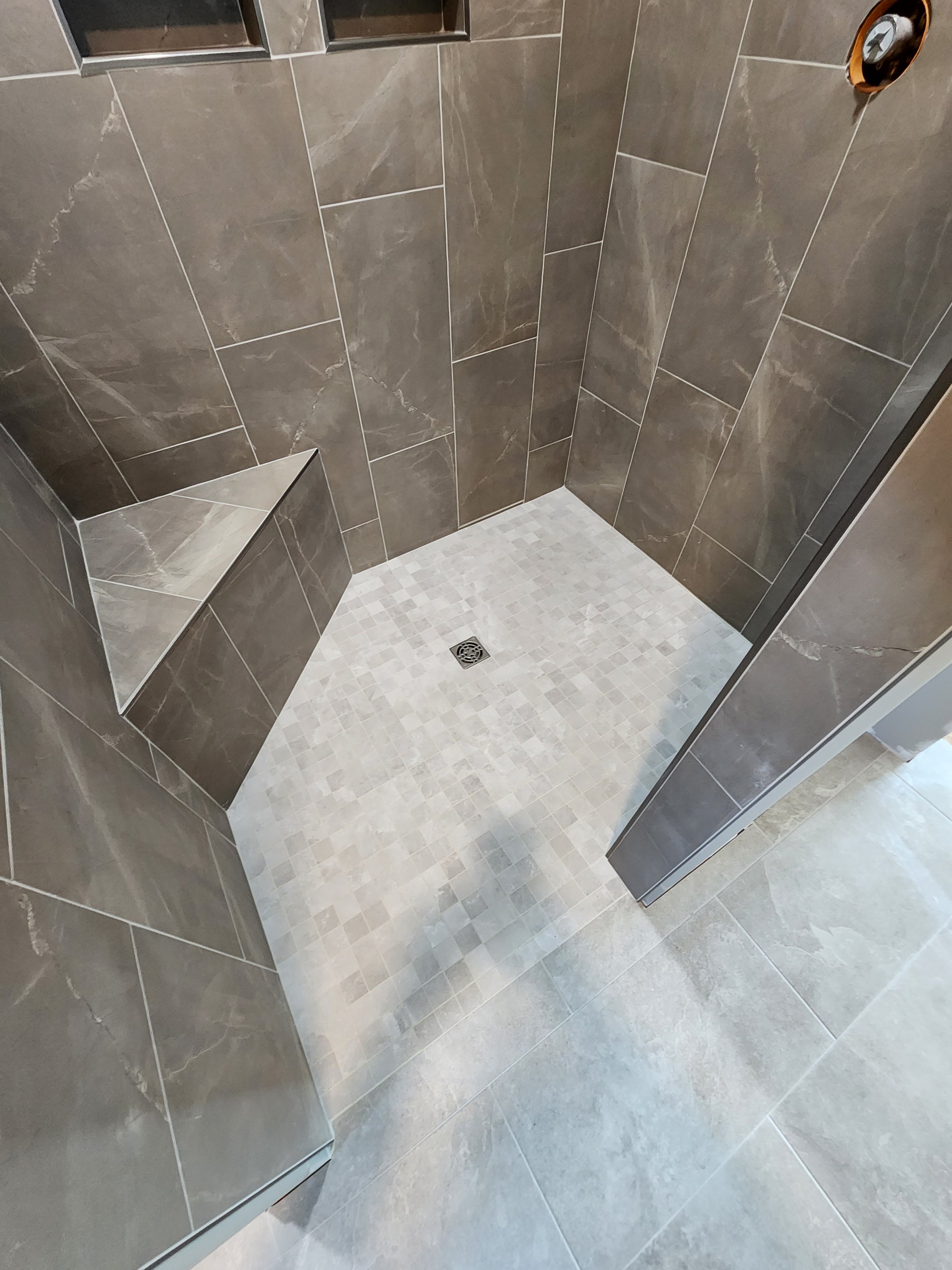 A modern shower with large brown marble wall tiles, small light beige square floor tiles, a built-in corner bench, a square floor drain, and a partial view of a clear glass shower door installed by Fourth Gen Flooring in Cedar Rapids Iowa.