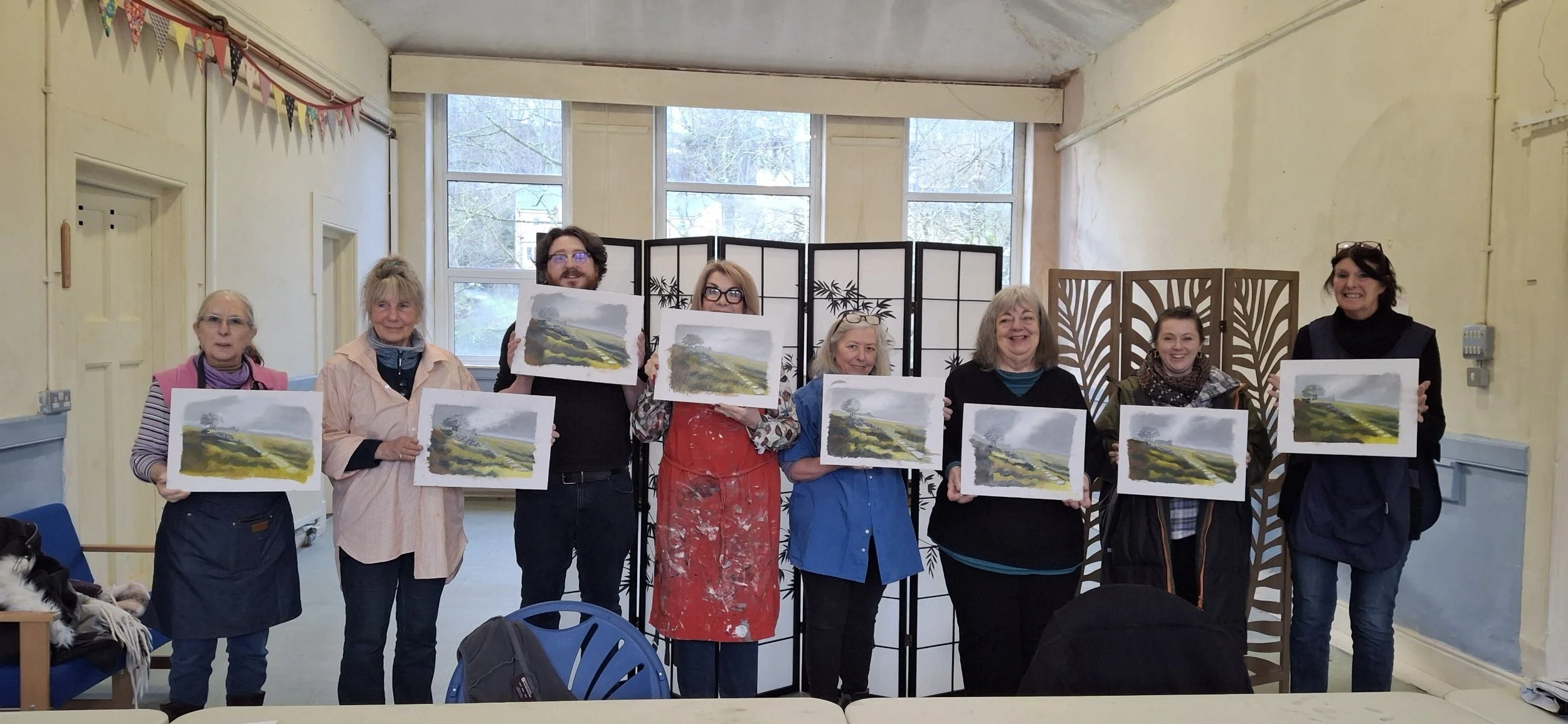 Group of eight people standing in a room, each holding a watercolor painting of a landscape with green hills and trees, with folding room dividers behind them.