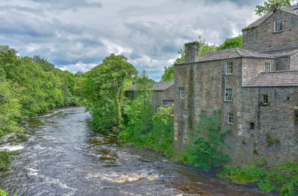 A scenic view of an old stone building by a river with lush green trees and cloudy sky in the background.