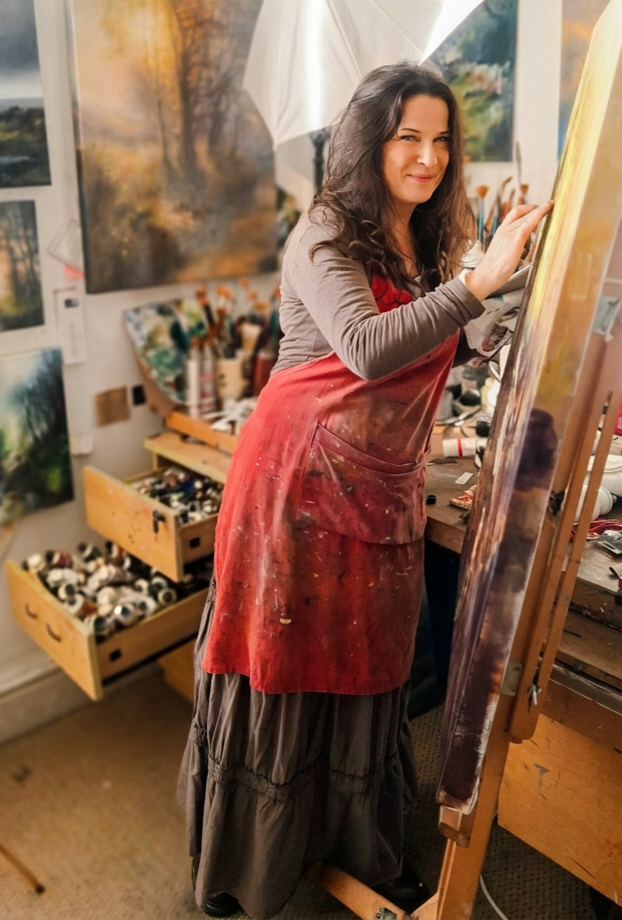 A Female artist in an art studio, wearing a red apron, standing and working on a painting on an easel with a smile.