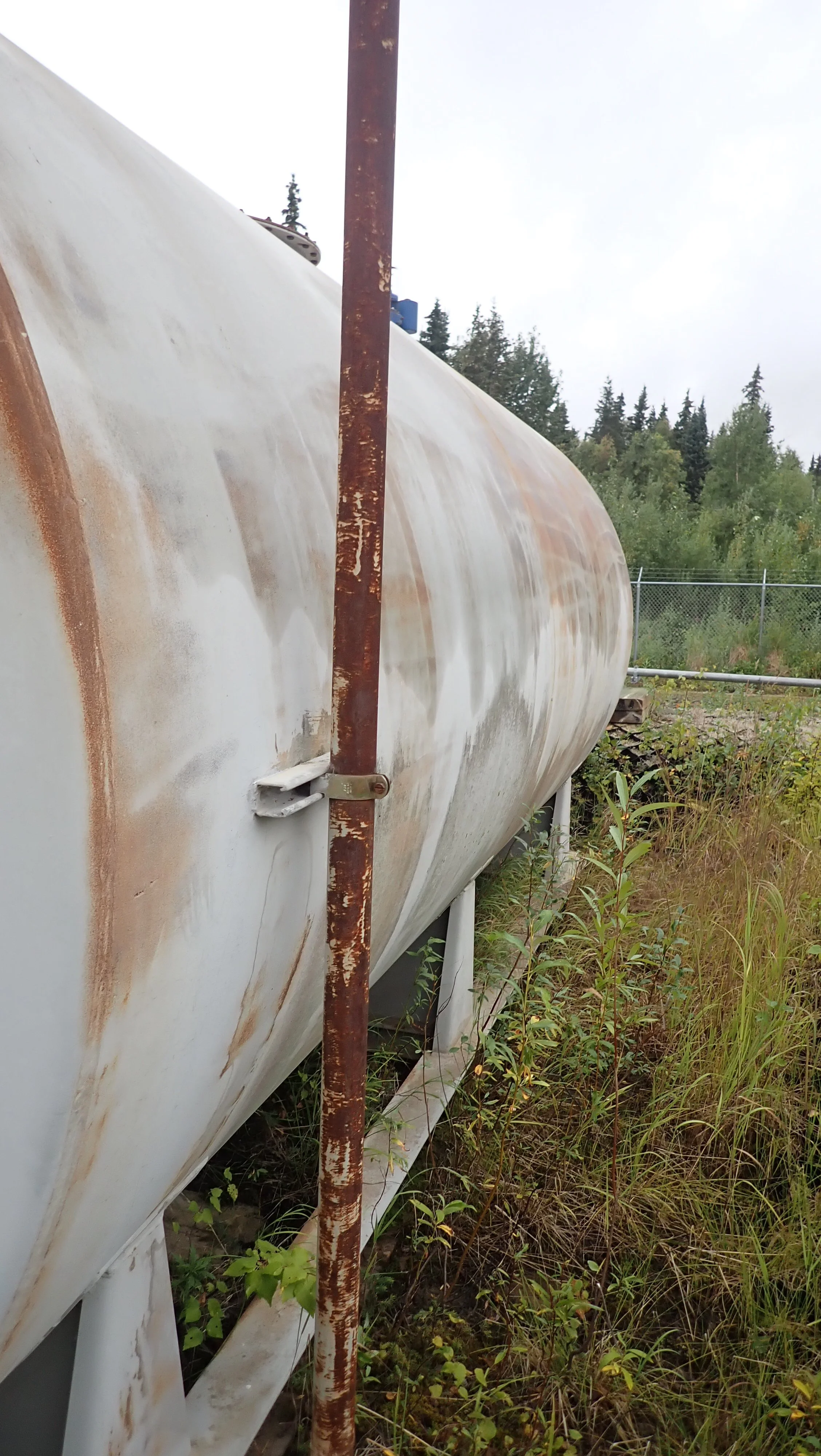 Horizontal fuel storage tank with surface rust and surrounding vegetation.
