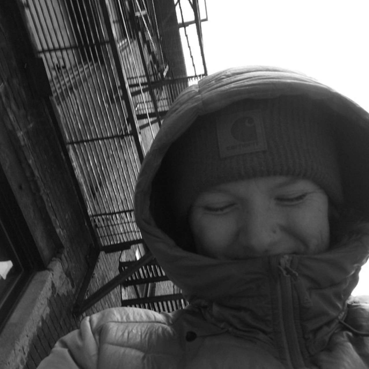 A person wearing a Carhartt beanie hat and a winter jacket, smiling with eyes closed, standing outdoors near a metal fence structure.