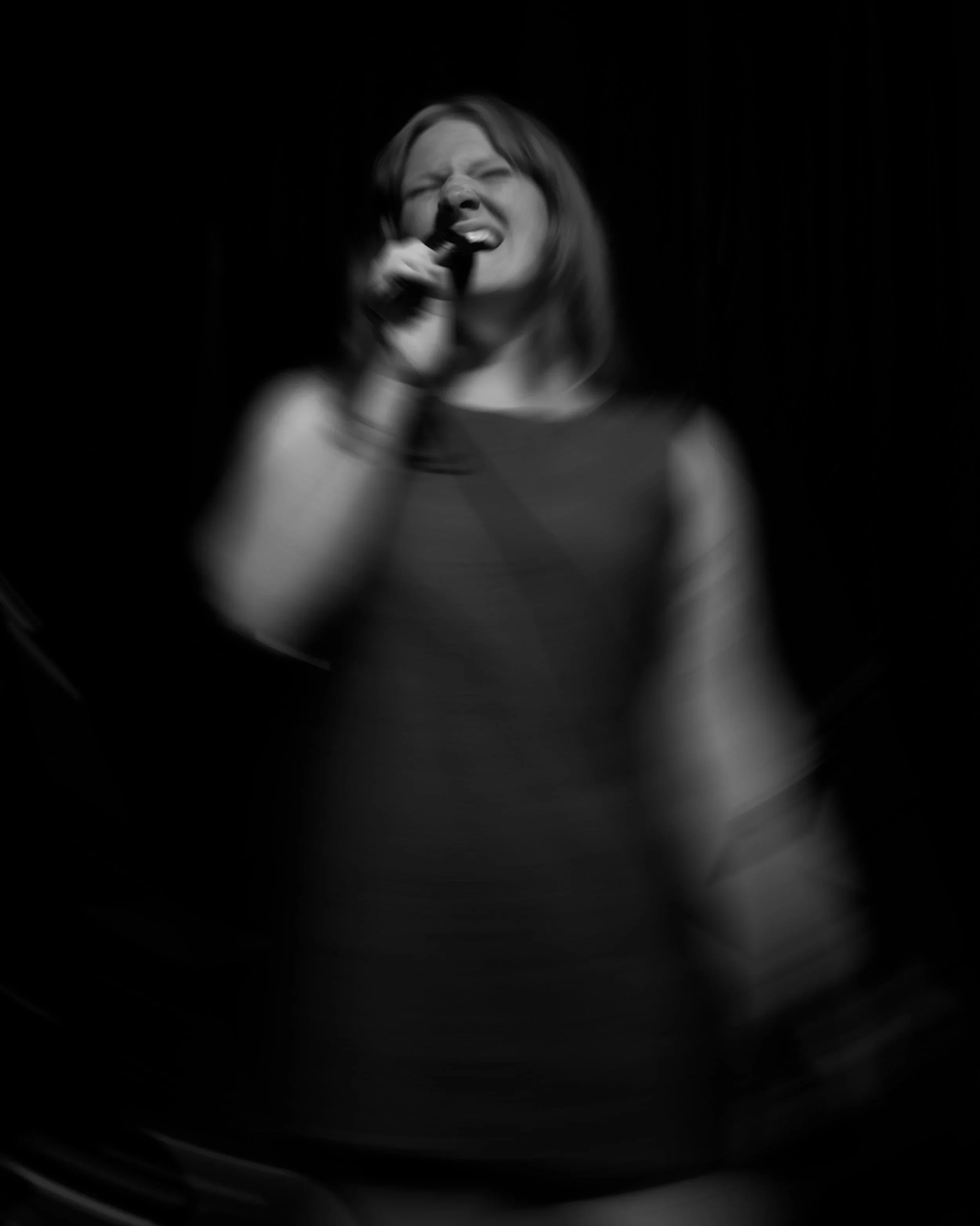 Black and white photo of a woman singing, holding a microphone with an emotional expression, against a dark background.