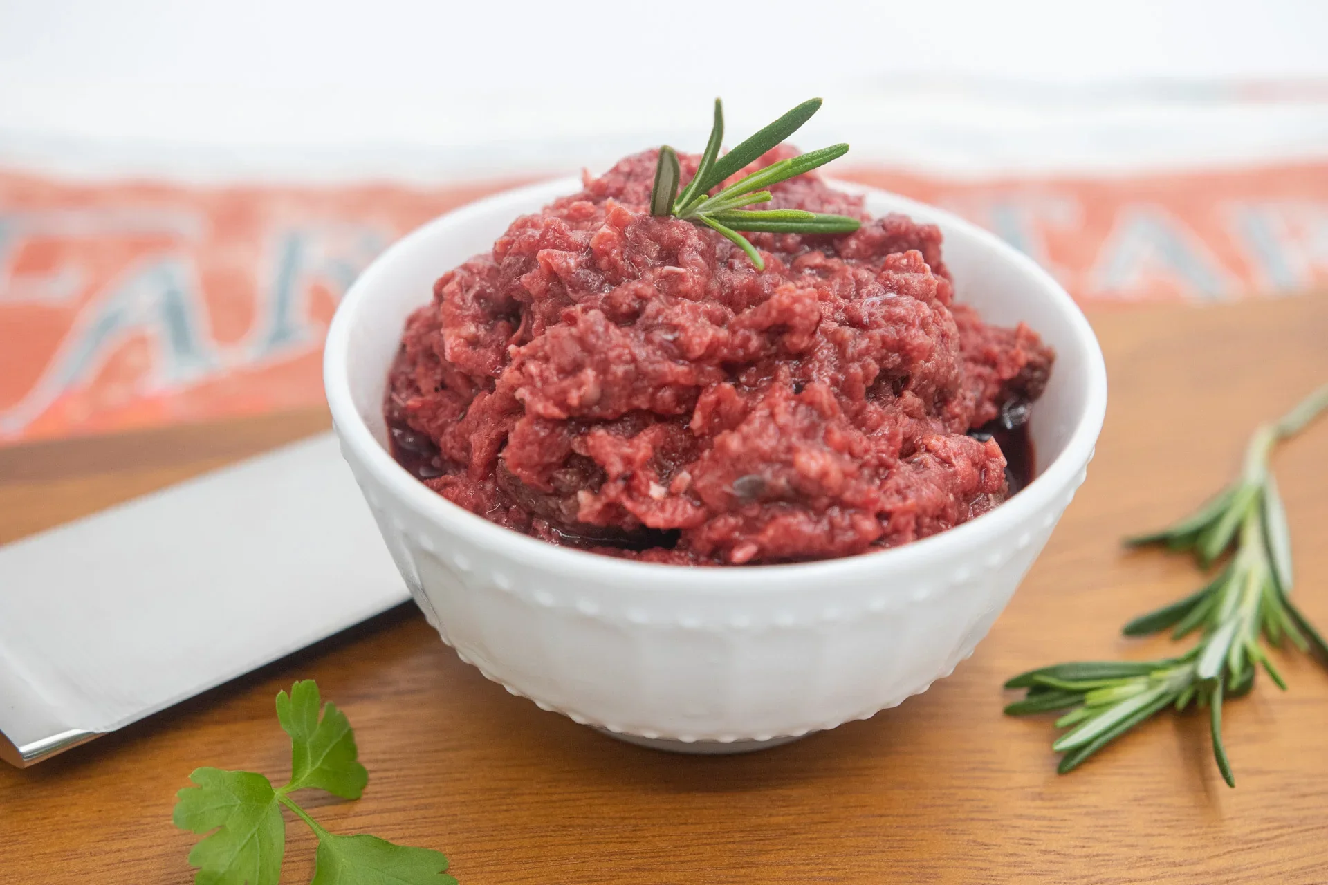 A white bowl of red-colored pet food garnished with rosemary on a wooden surface.