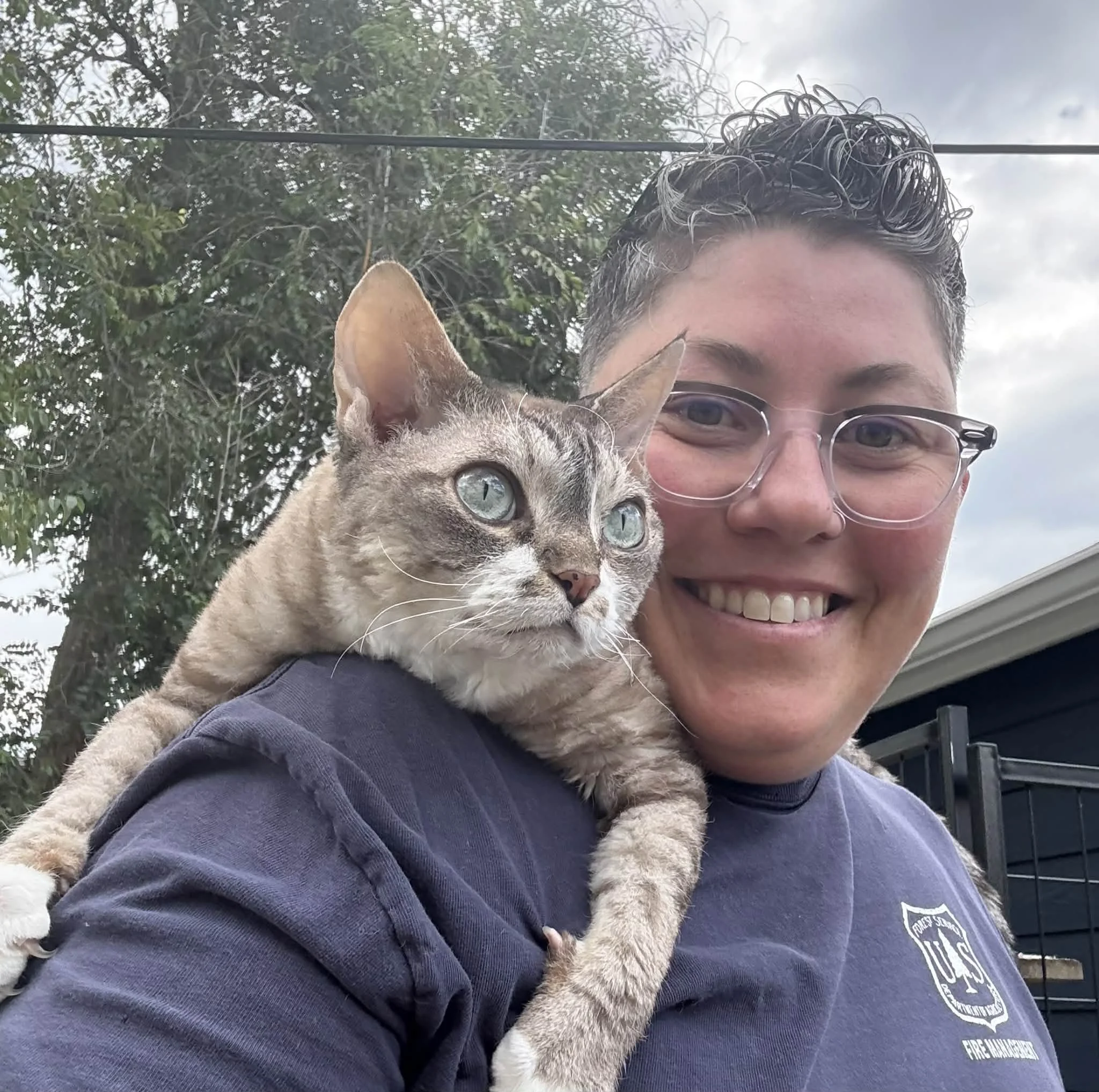 A person with short curly hair, glasses, and a navy blue shirt is smiling while holding a gray tabby cat on their shoulder outdoors. The background includes a tree and a cloudy sky.