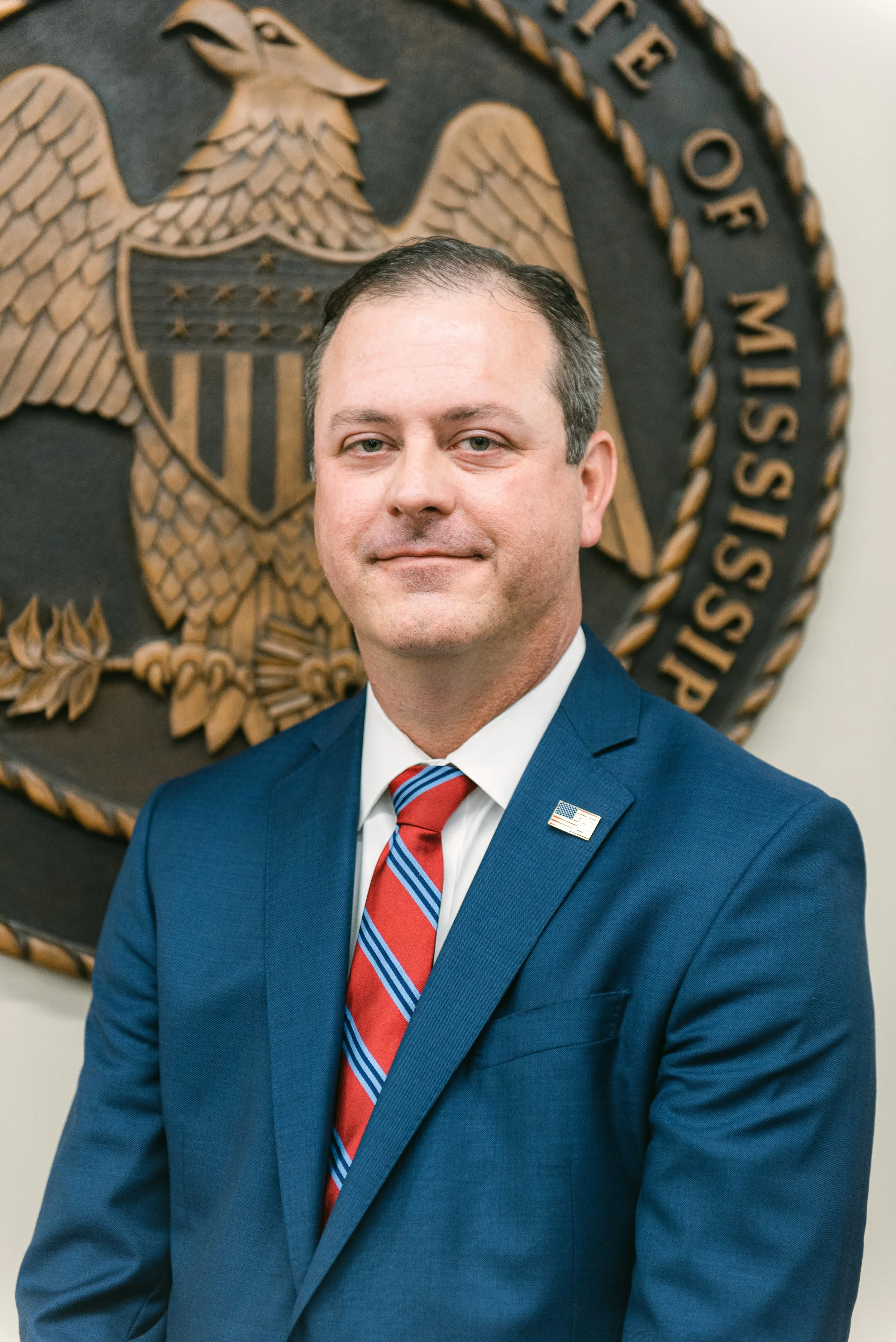 A man in a blue suit with a red, white, and blue striped tie, standing in front of a wall with the U.S. Department of Justice emblem.