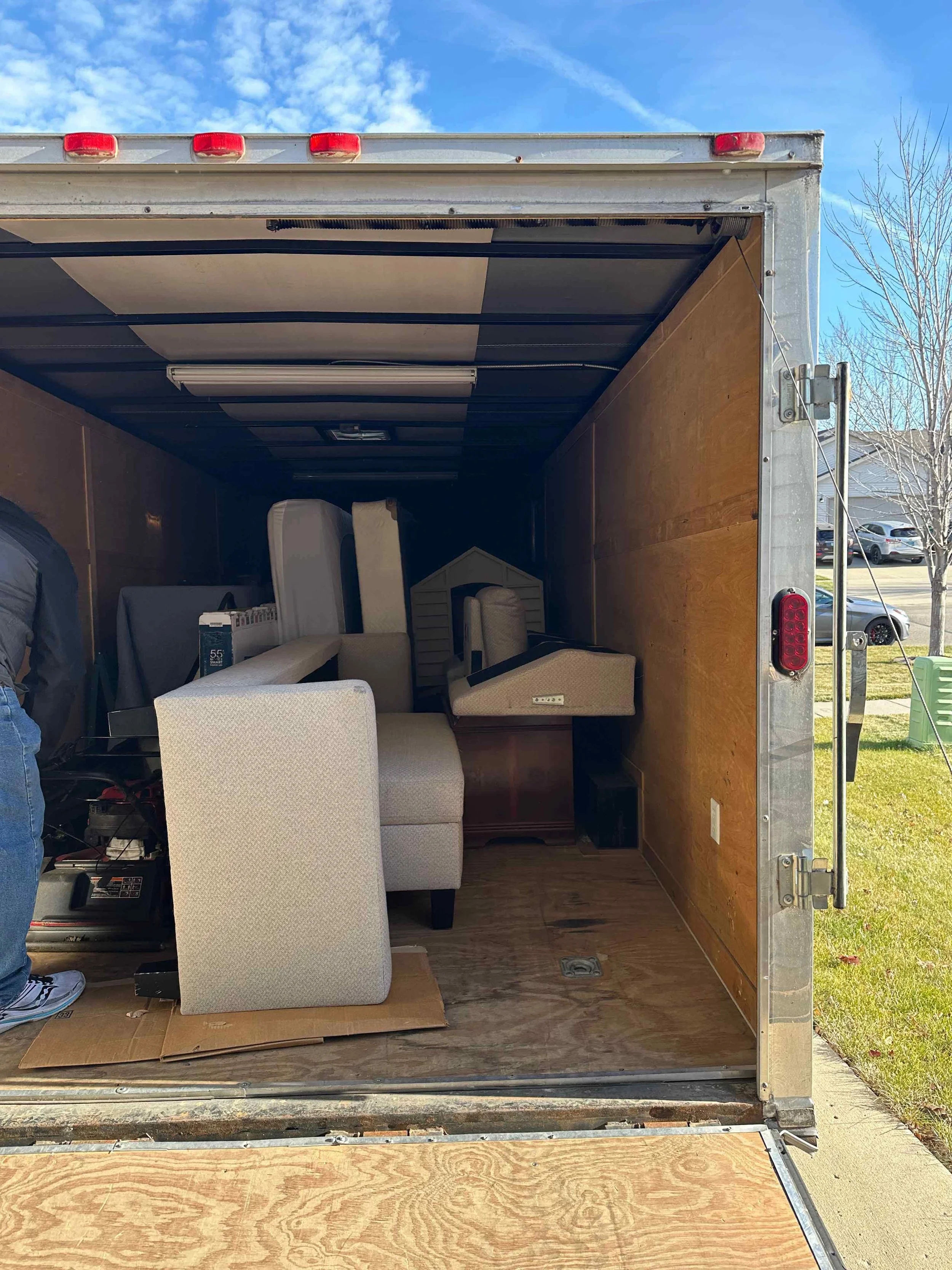 The back of a moving truck filled with furniture, including a beige armchair, a small wooden table, and a plastic storage shed, with a person partially visible on the left side.