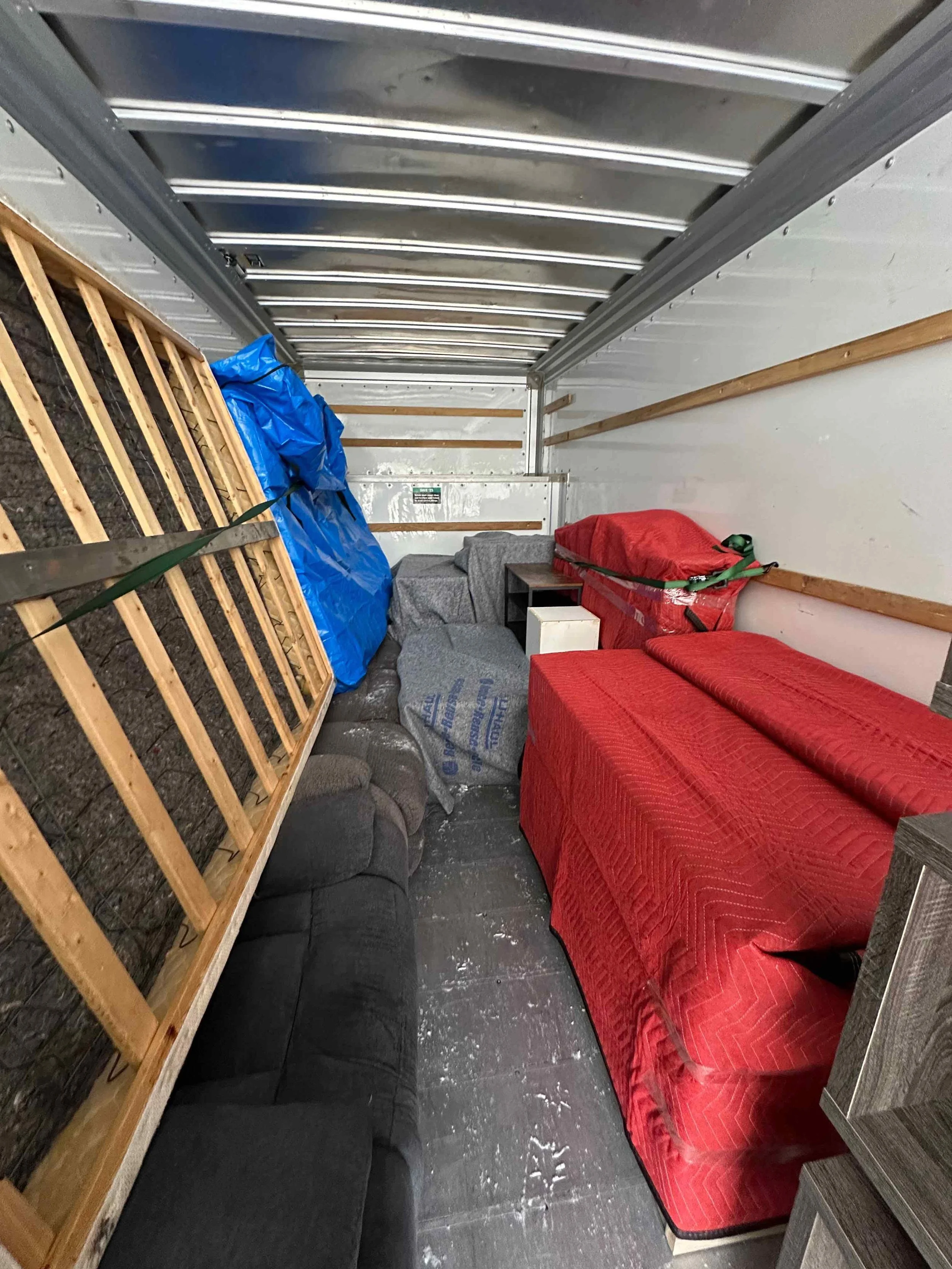 The inside of a storage unit with various furniture and items, including a gray couch, a red covered couch, a small white cabinet, a black table, and a mattress wrapped in plastic, with some pallets and blue tarp on the side.