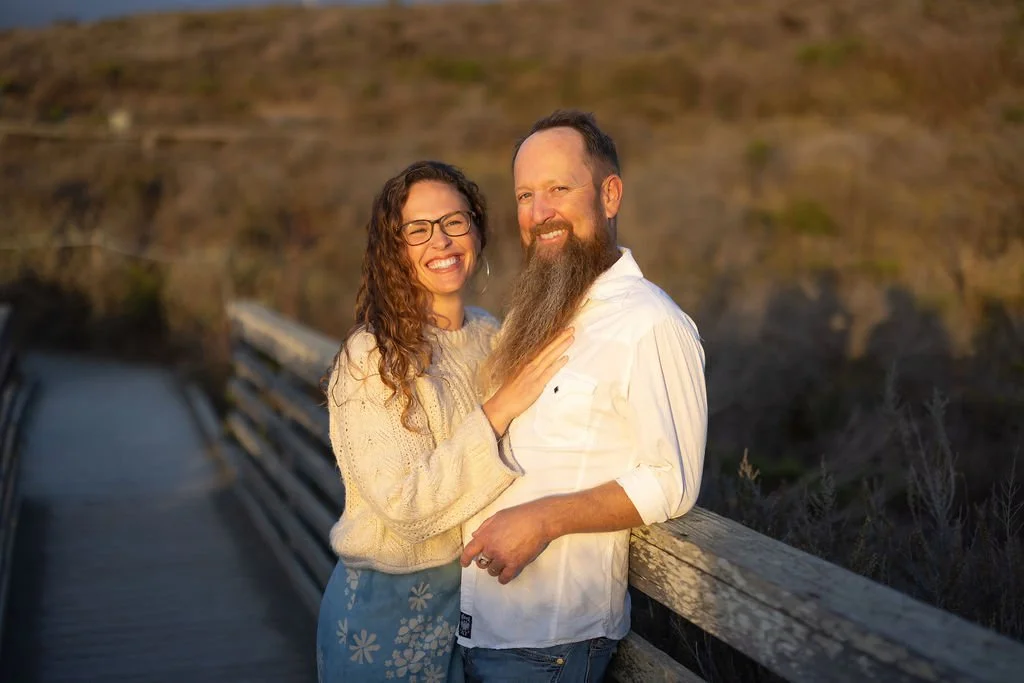 A smiling woman with glasses and curly hair standing next to a bearded man on a wooden bridge outdoors during sunset.