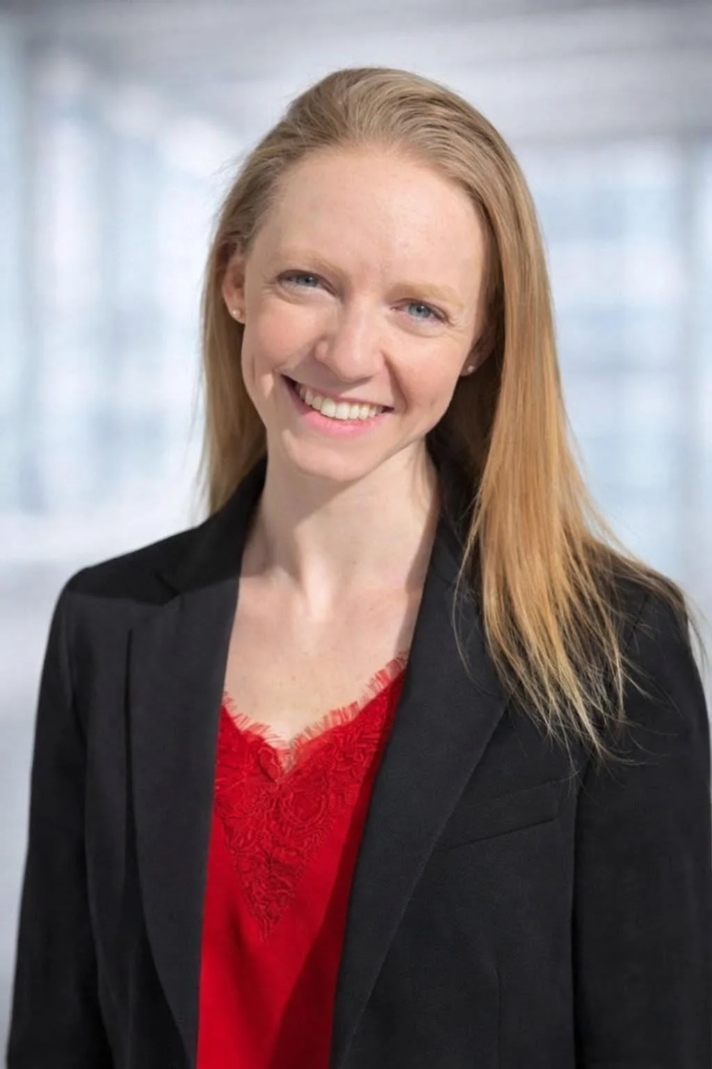 A woman with long red hair, wearing a black blazer and red top, smiling at the camera with a blurred office background.