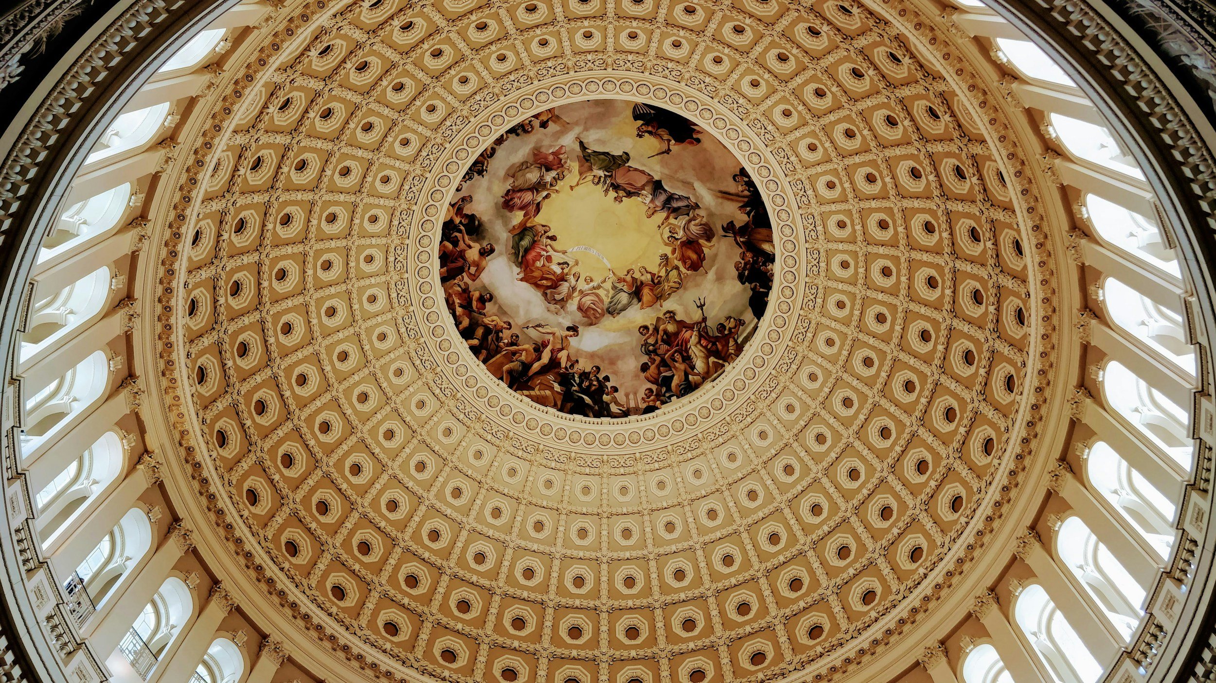 The ornate domed ceiling of the U.S. Capitol building with a detailed painting surrounded by intricate architectural patterns and columns.