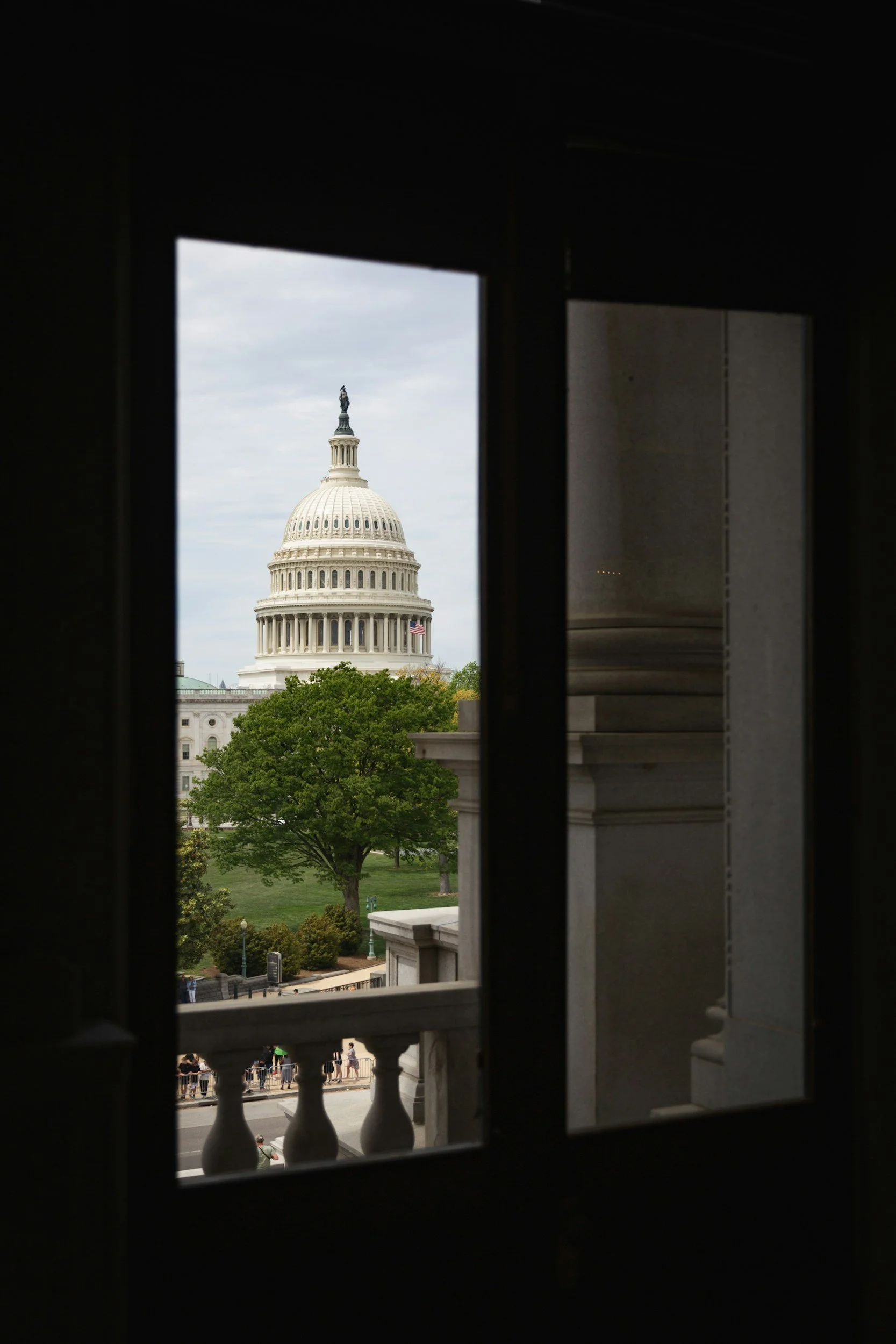 View of the U.S. Capitol building through a window with dark framing, showing the dome and a partly cloudy sky.