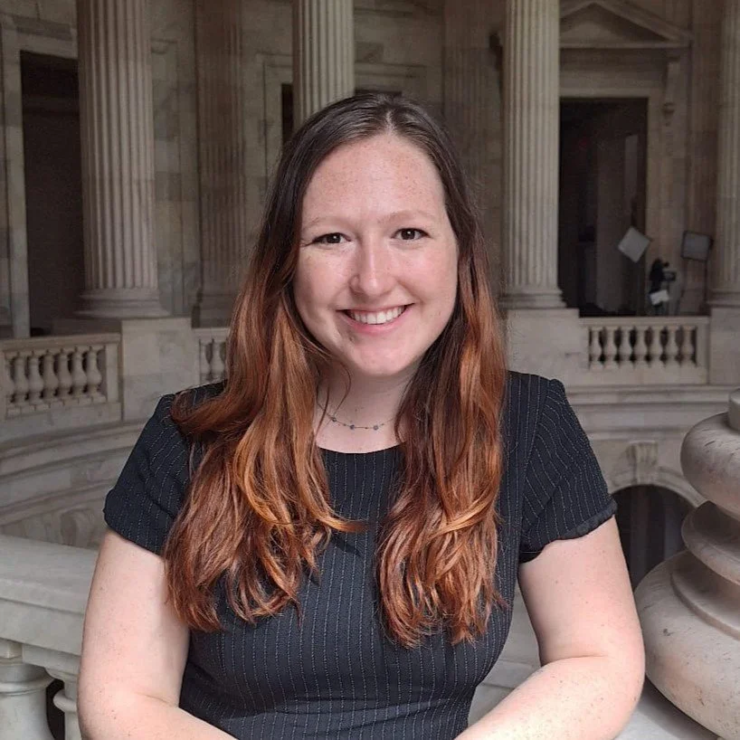 A young woman with long, wavy, red hair smiling and posing inside a historic building with columns and ornate architecture.
