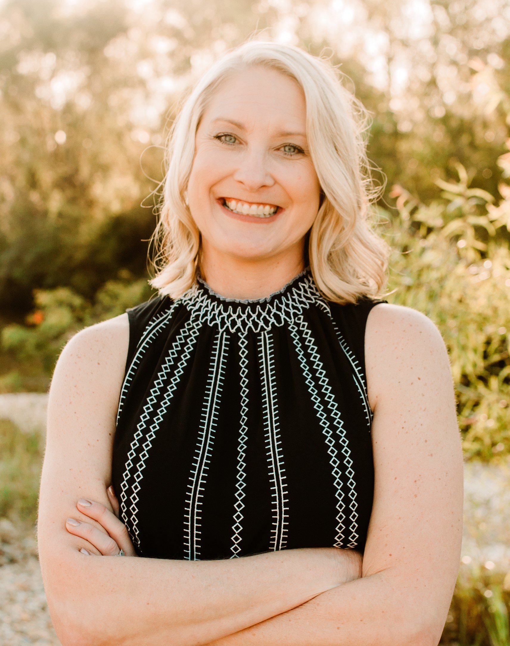 A woman with blonde hair smiling outdoors in front of greenery, wearing a sleeveless black top with white embroidery details.