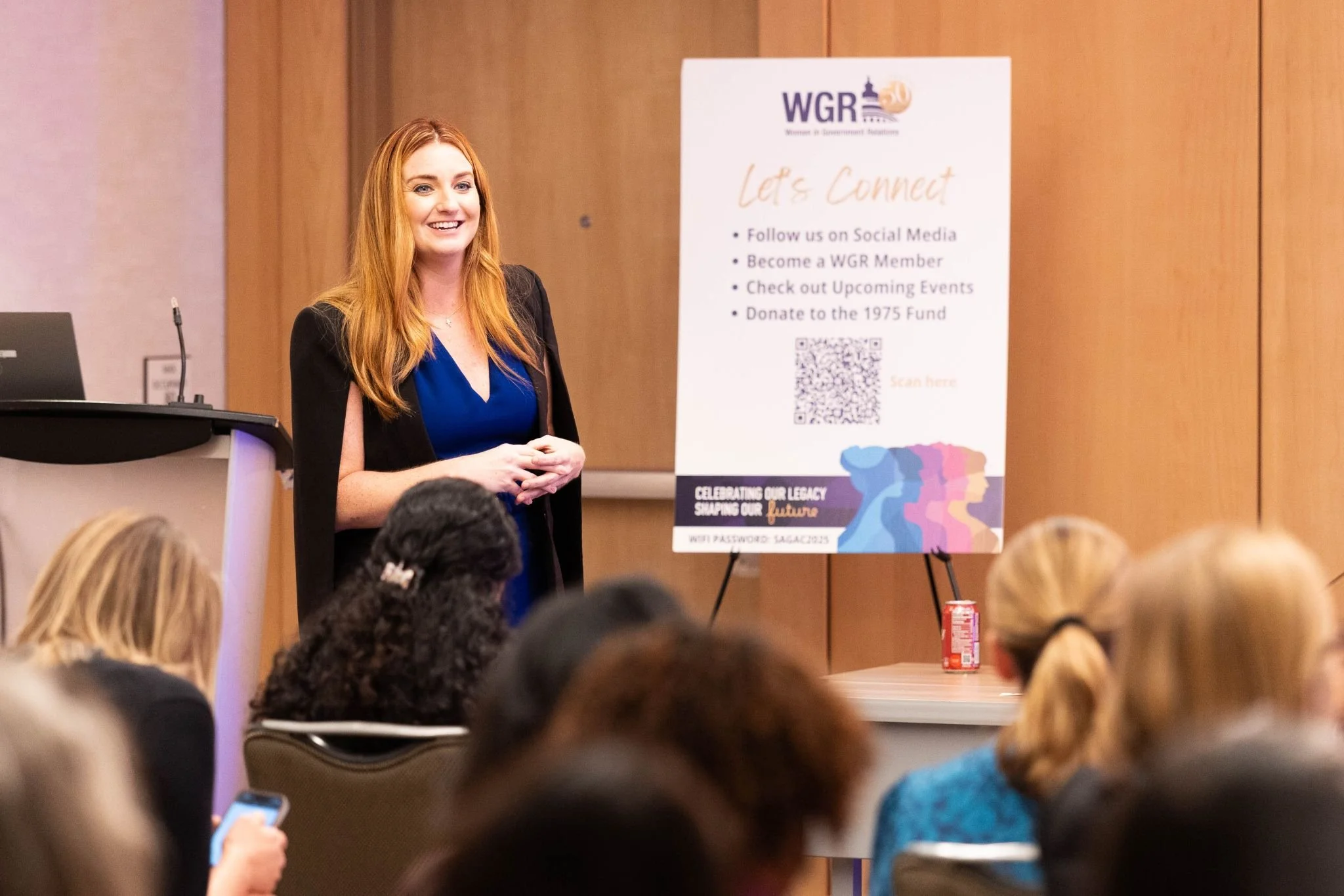 A woman with long red hair wearing a blue dress and black jacket stands in front of an audience at a conference, smiling. Behind her is a white poster board on a stand with blue and pink graphics and text about connecting, social media, and donation opportunities.